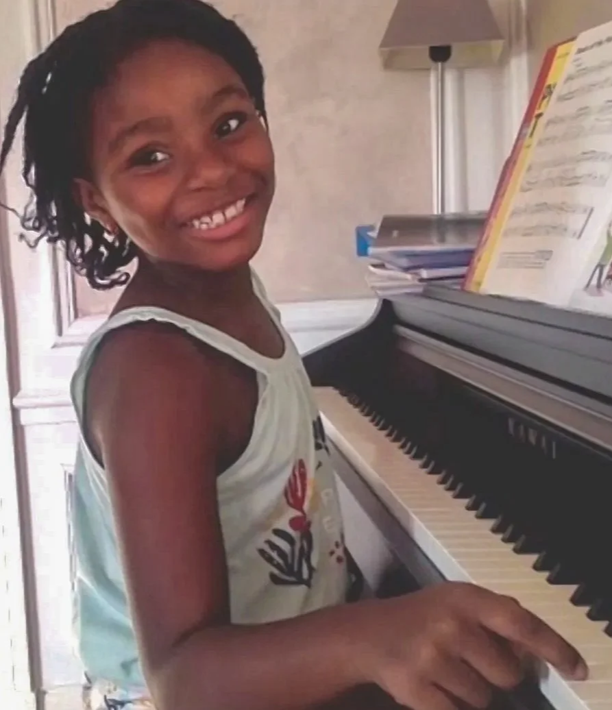 A young girl with a joyful expression playing a piano, with sheet music on the music stand, in a home setting.