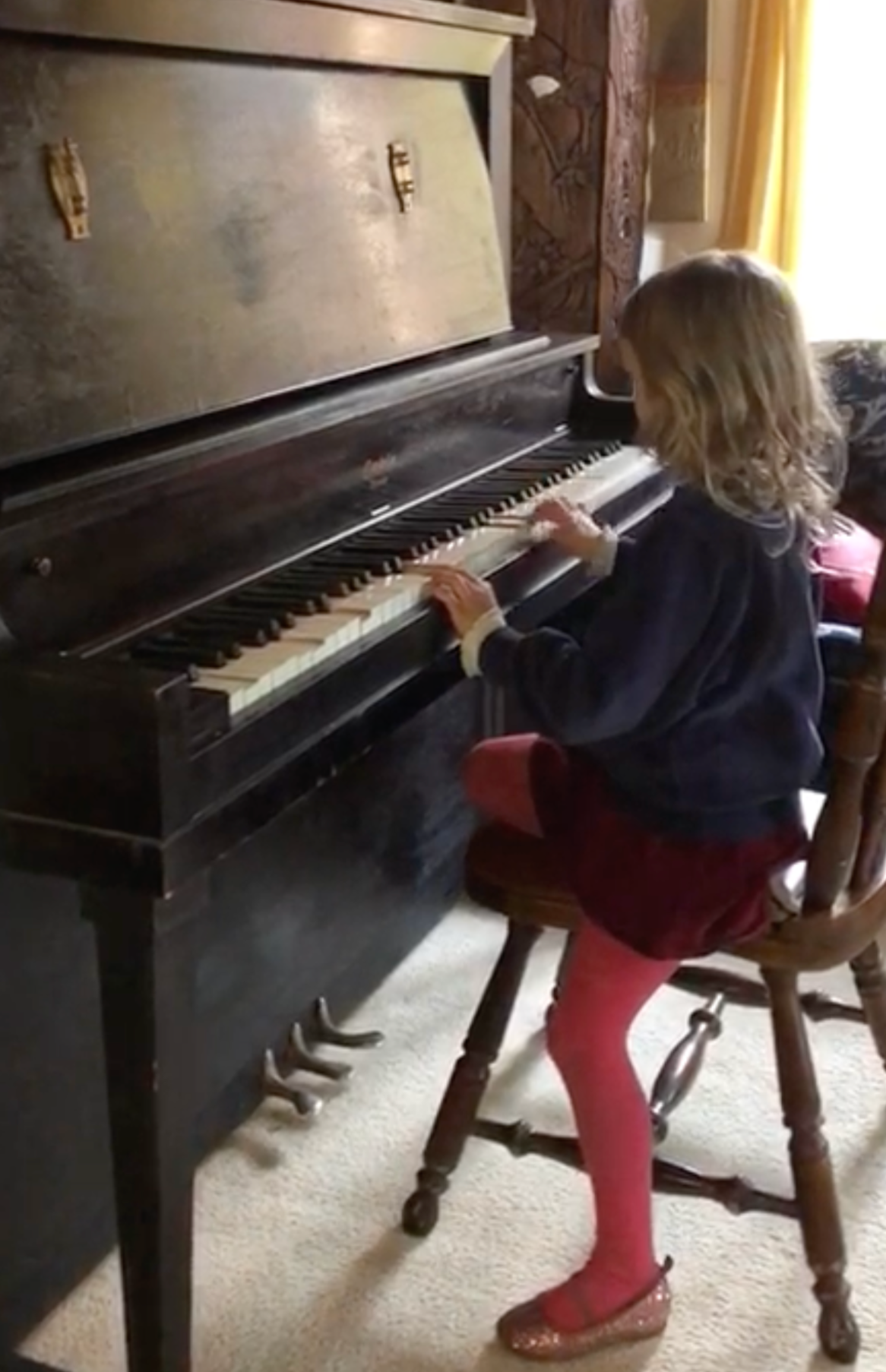 A young girl with blonde hair playing an upright piano in a cozy room with yellow curtains and a Persian-style rug.