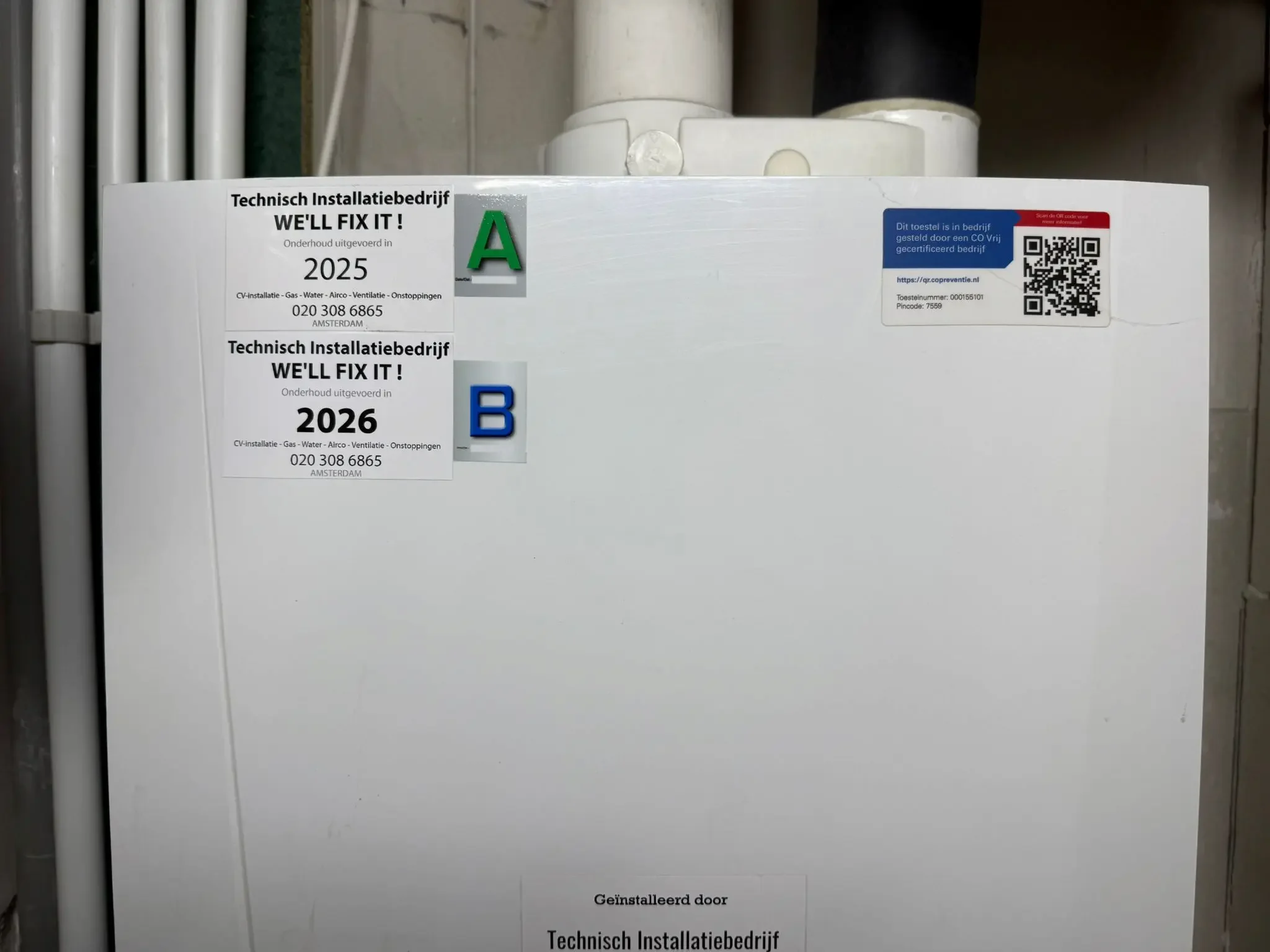 A white utility box with labels indicating maintenance dates for 2025 and 2026, with a QR code and technical information in Dutch, and labeled with letters A and B.