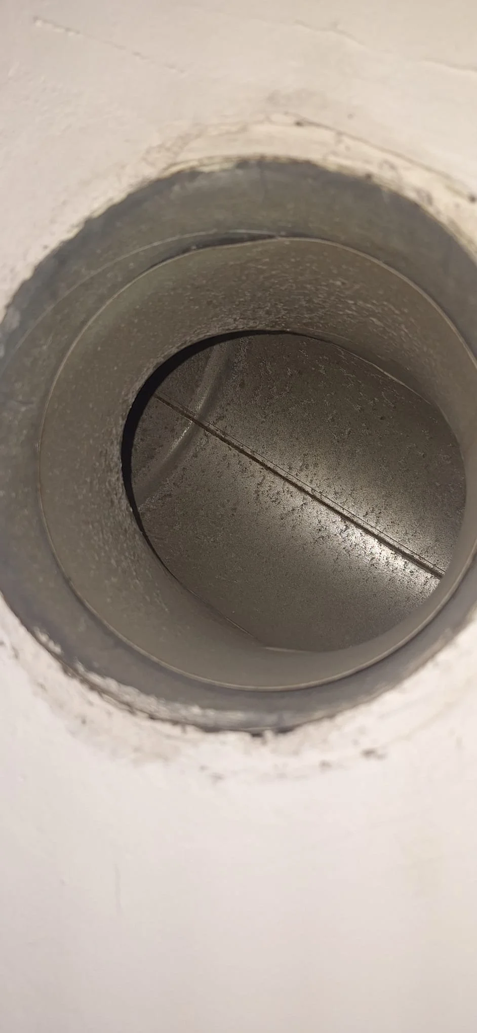 Close-up view of a metallic pipe or vent in a wall, showing a cylindrical shape with some dust and dirt buildup inside.
