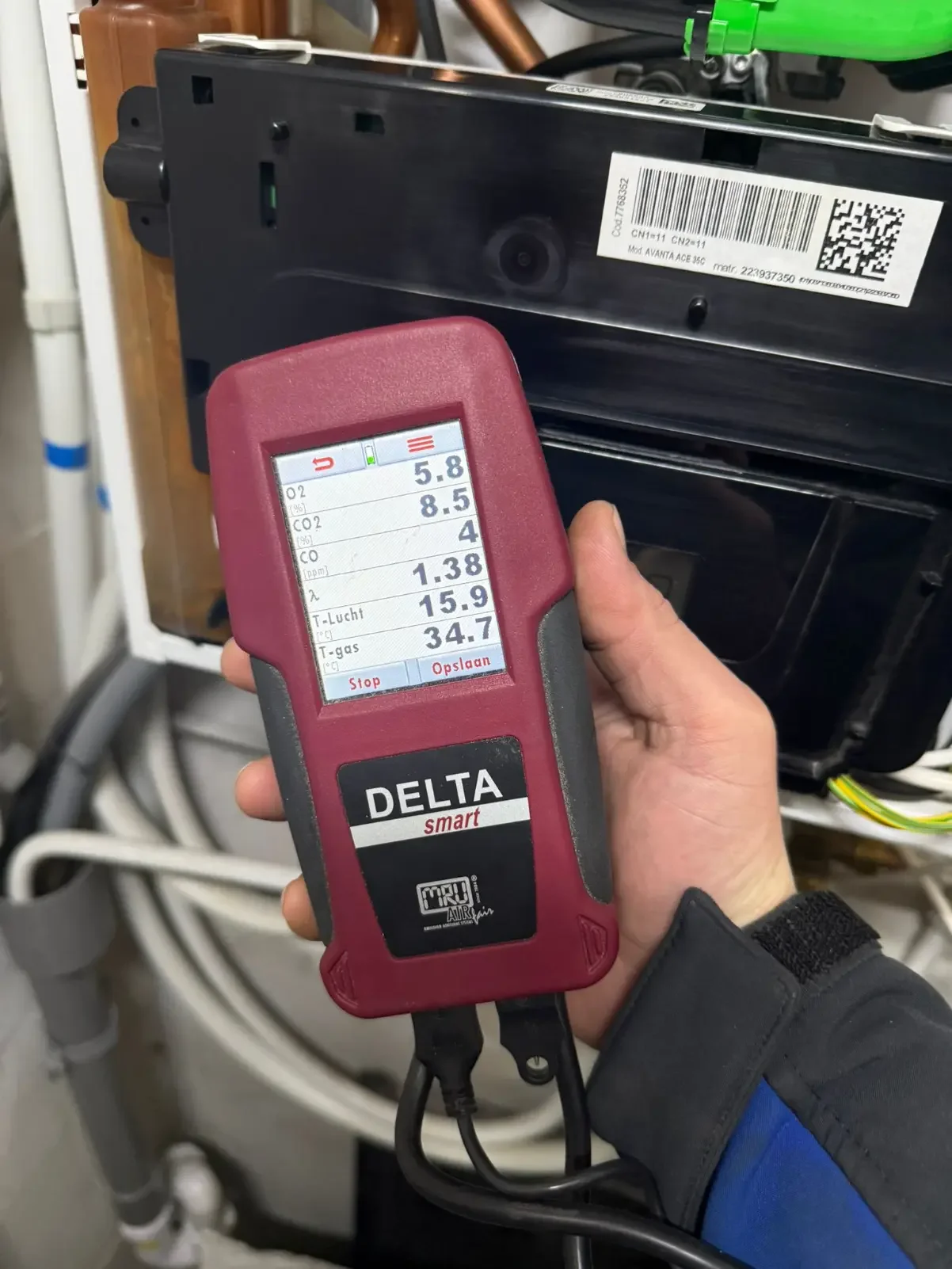 Hand holding a Delta smart air quality meter in front of an HVAC system, displaying measurements for oxygen, carbon dioxide, carbon monoxide, nitrogen, temperature, and humidity.