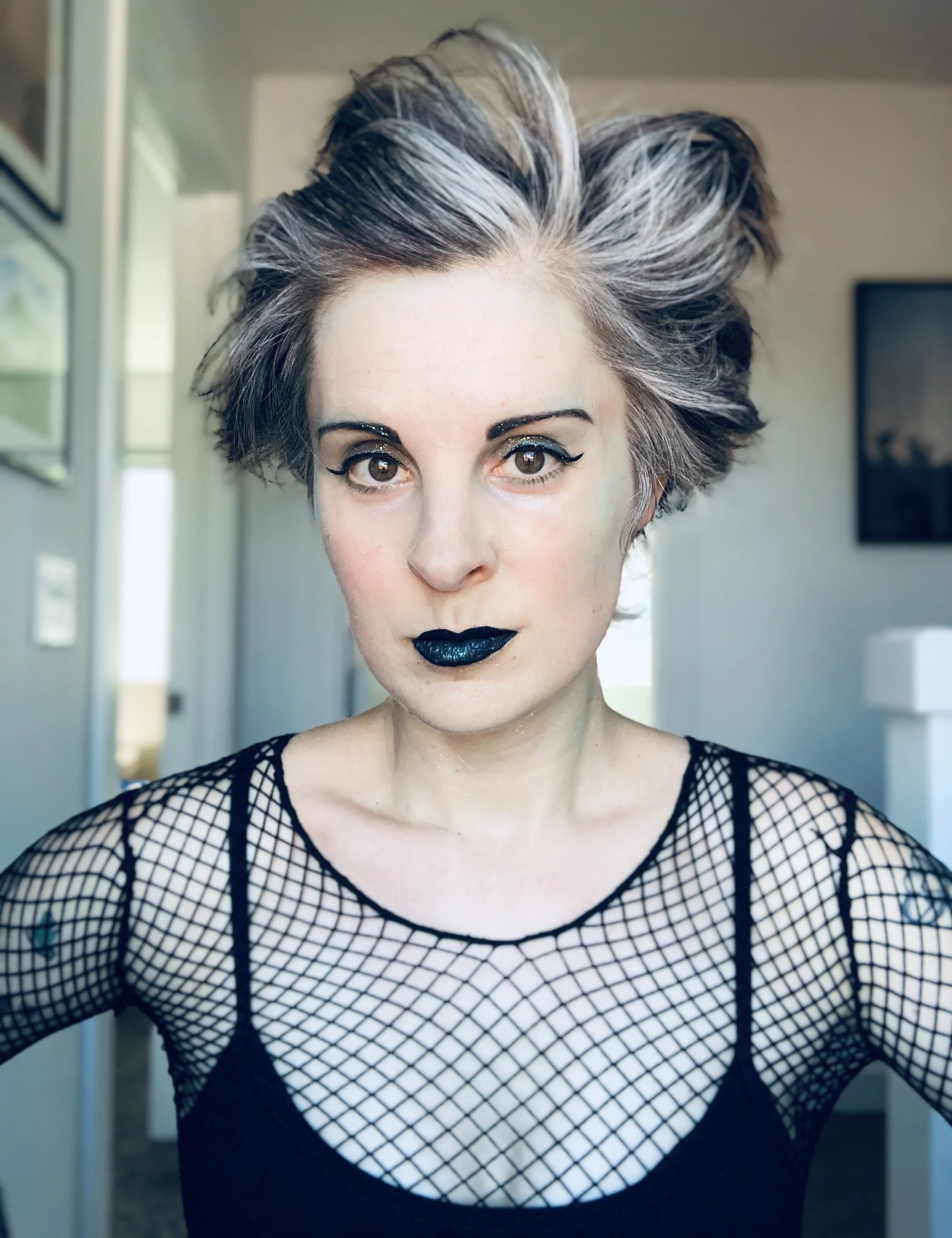 Selfie of Jen from 2020, she's wearing a black sport bra, black fishnet top, black pixie cut growing out with grey streaks and styled up with dramatic makeup. 