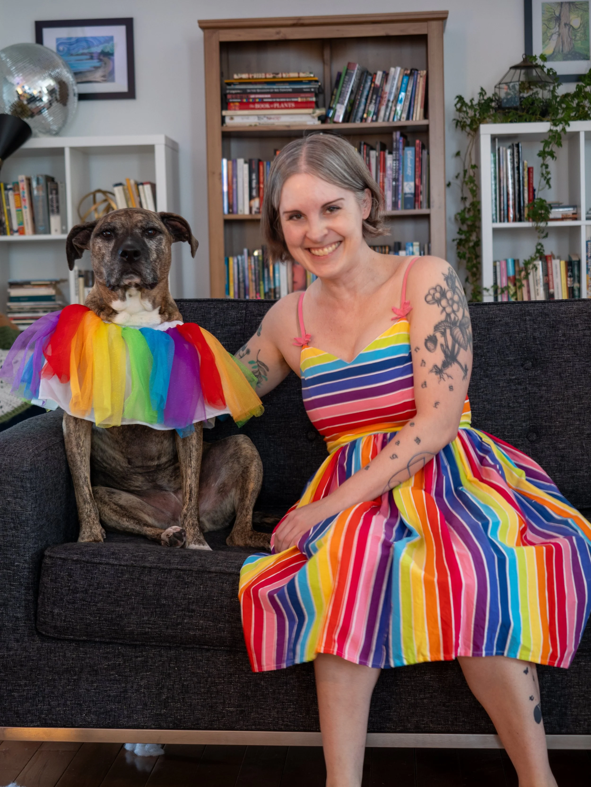 Photo of jen from 2025 taken by Jeff Woodward, she's wearing a rainbow dress and sitting on the couch and Dolly the brindle pittbull is next to her wearing a rainbow tutu as a cape.