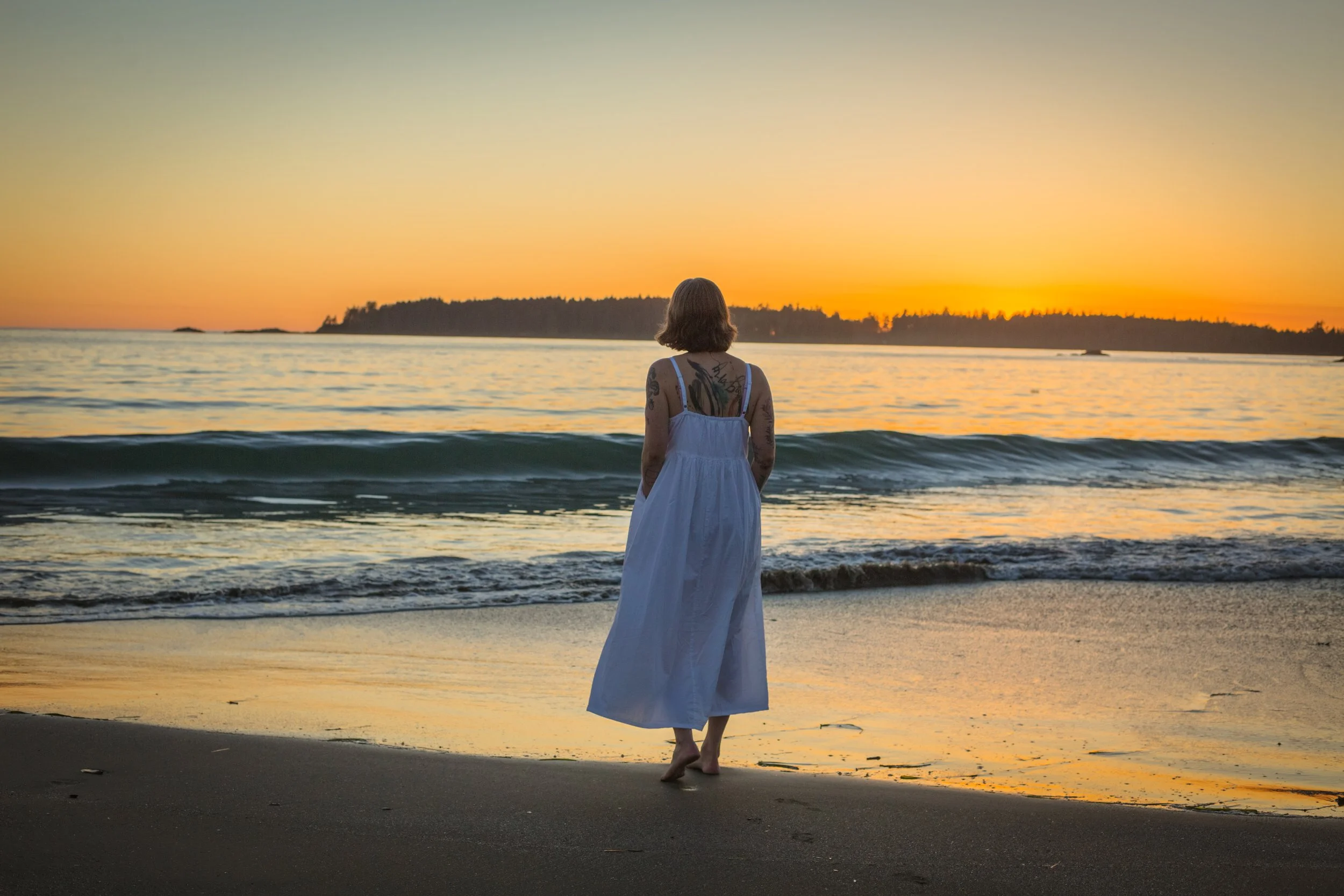 Photo of Jen taken by Jeff Woodward in September 2022, sunset on Mackenzie Beach in Tofino with Jen wearing a white dress and walking towards the ocean.