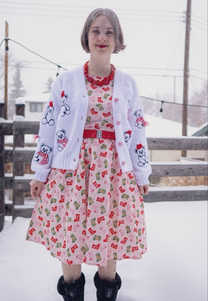 Photo of Jen from 2025 taken by Jeff woodward where she's standing outside in the snow wearing a pink dress with red and green christmas stockings, red belt, red button necklace, and white sweater with teddy bears that have little scarves and hats.