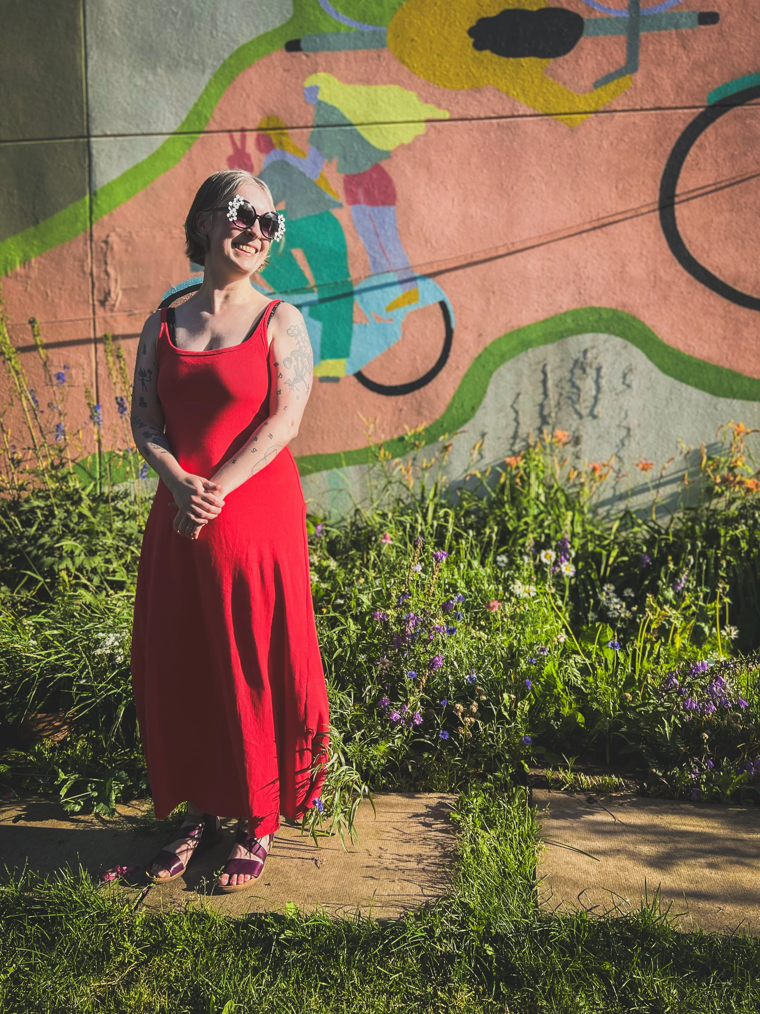 Photo of Jen from 2025 taken by Jeff Woodward  standing in the garden at Otto restaurant wearing a. red dress with a bike mural behind her.