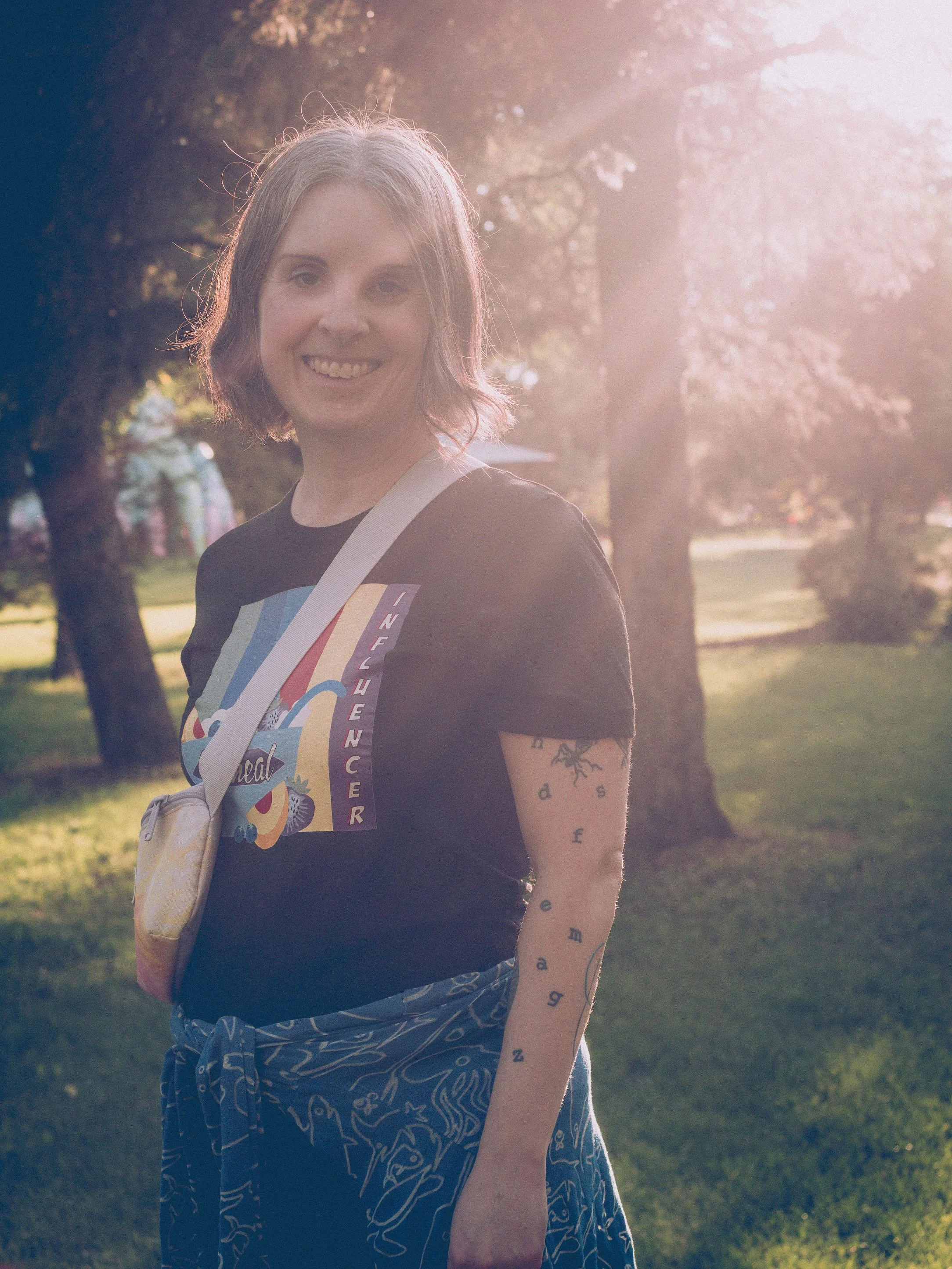 Photo of jen from 2025 taken by Jeff Woodward standing in a sunbeans at Borden Park wearing a oatmeal influencer tshirt and fanny pack with yoga pants.