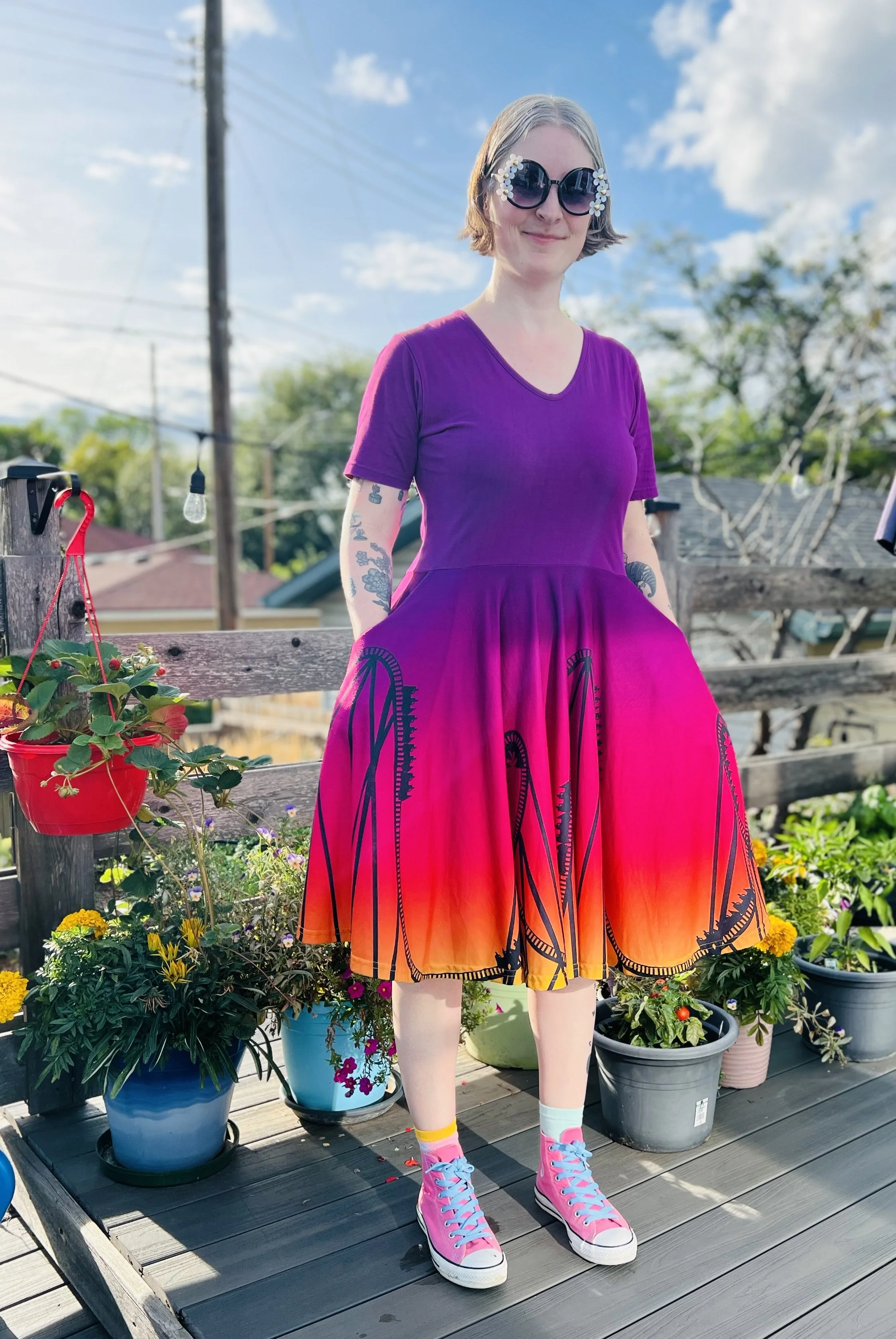 Photo of Jen from 2025 wearing a purple, pink and orange Svaha dress with black silhouette rollercoaster design, standing on their backyard deck next to flower pots.
