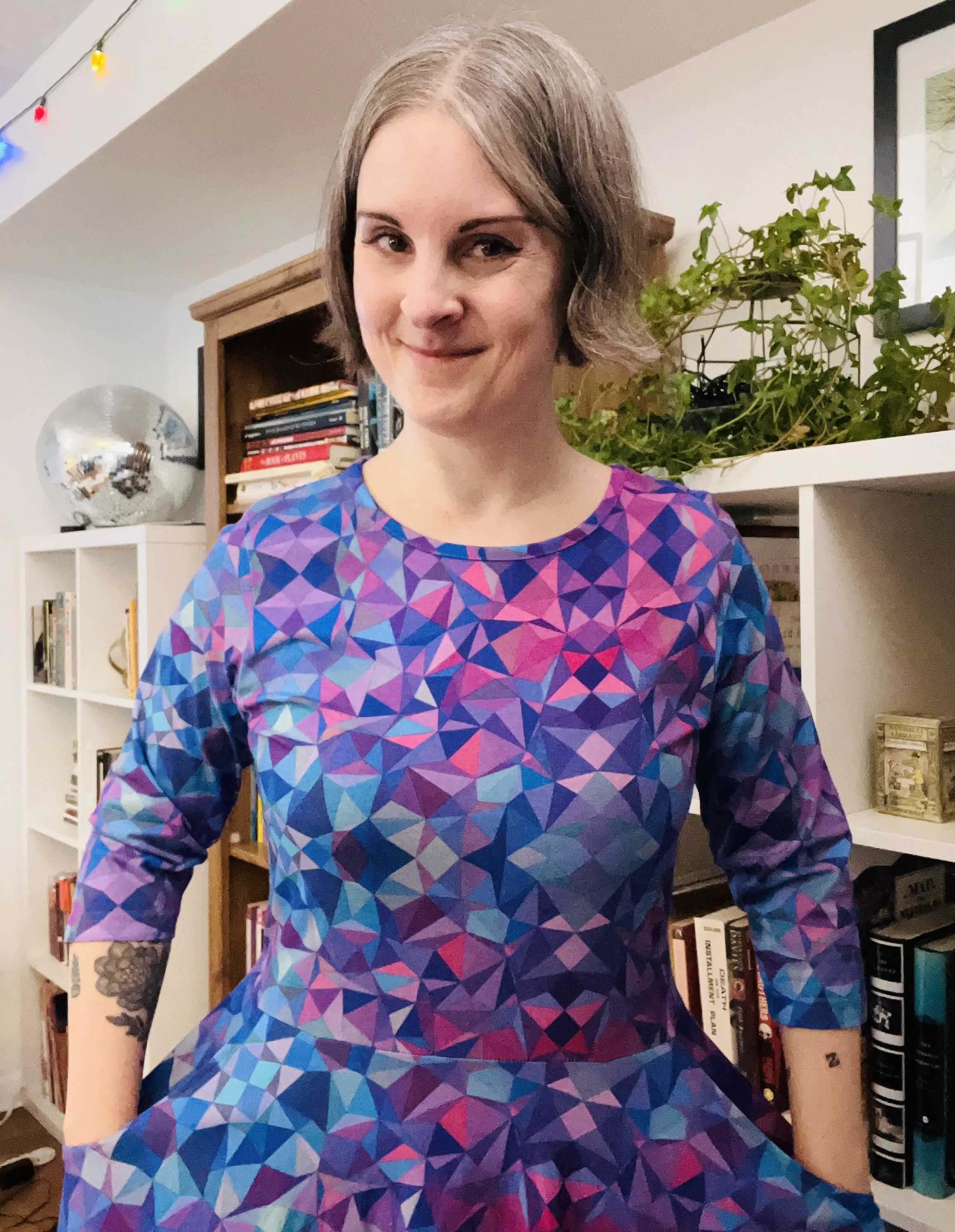 Selfie of Jen from 2026 standing in front of bookcases wearing a svaha dress with pink, blue and purple kaleidoscope pattern.