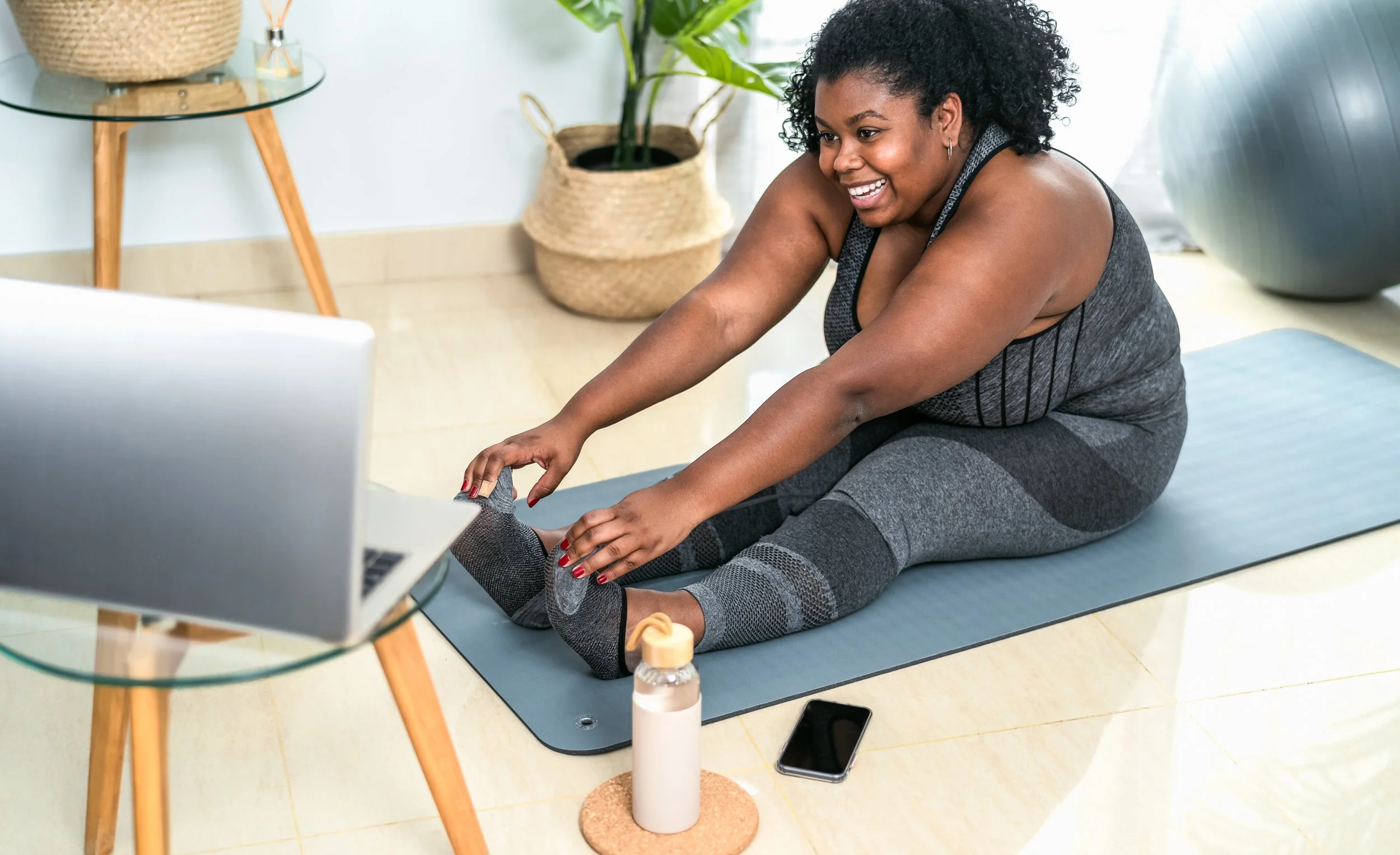 Woman in workout clothes stretching on a yoga mat during an online fitness class, with a laptop, water bottle, and smartphone nearby in a home gym setting.