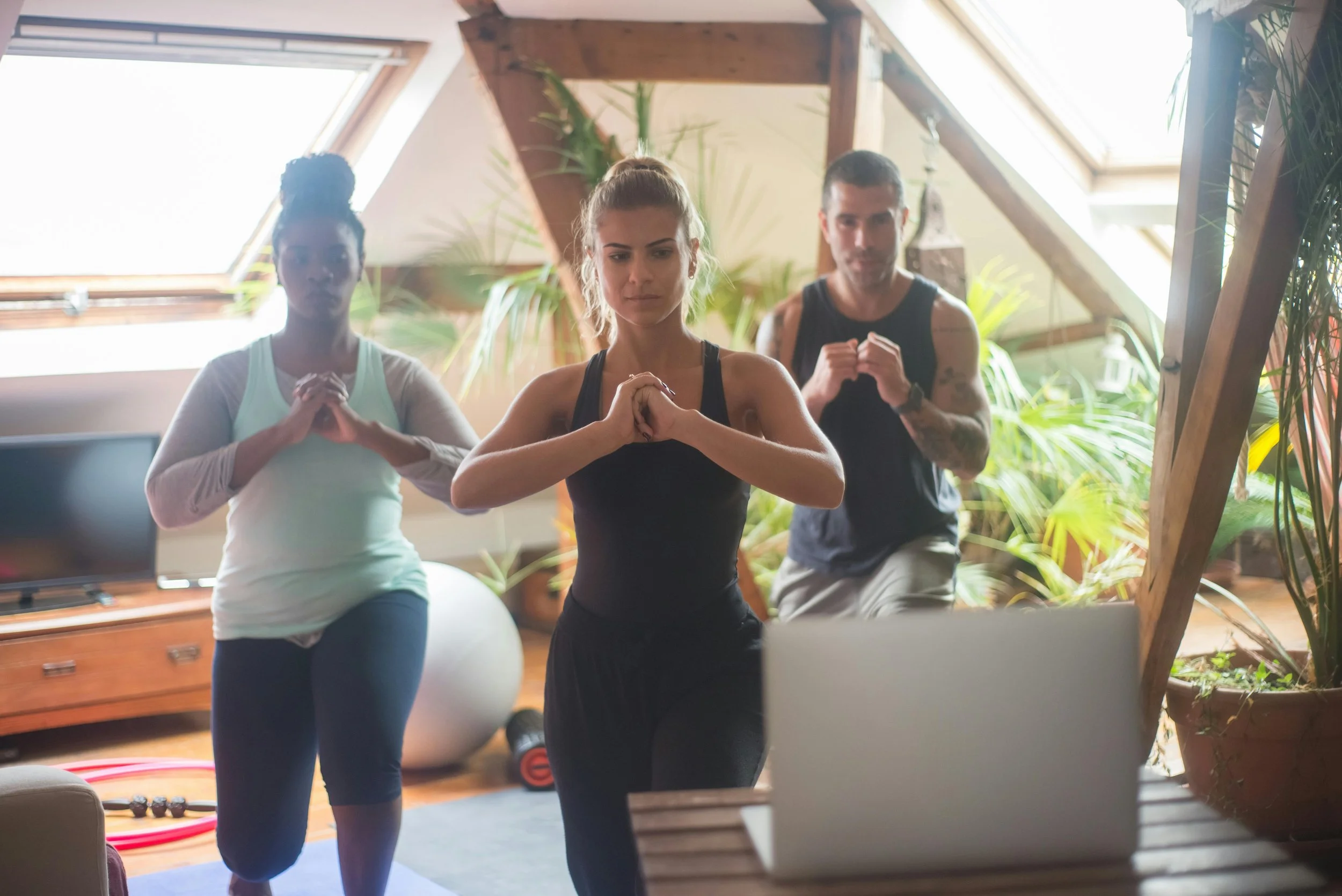 Three women are participating in a virtual yoga or fitness class in a living room with large skylight windows and abundant indoor plants.