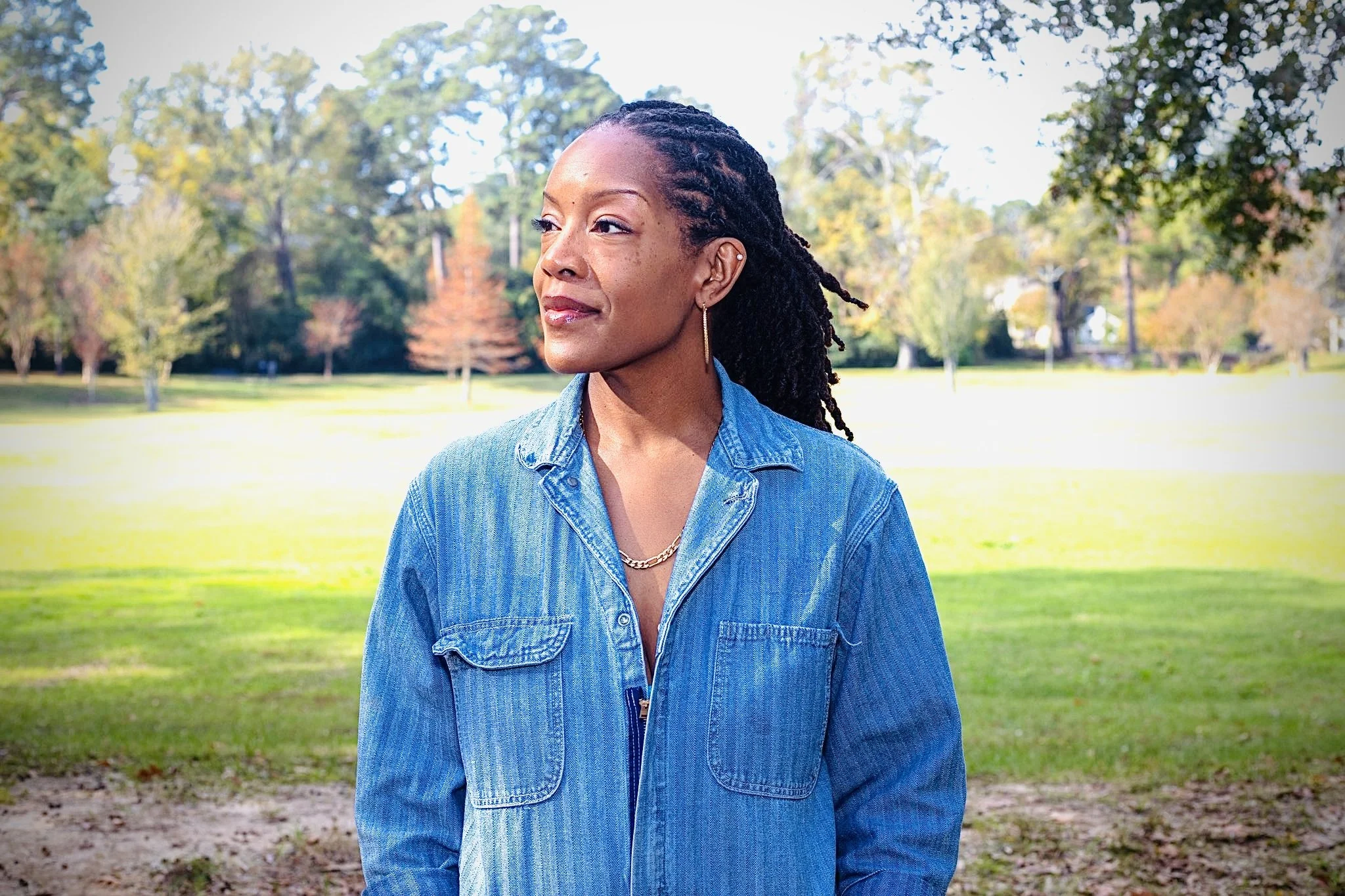 An African American woman with long dreadlocks, wearing a denim jacket, standing outdoors in a park with green grass and trees in the background.