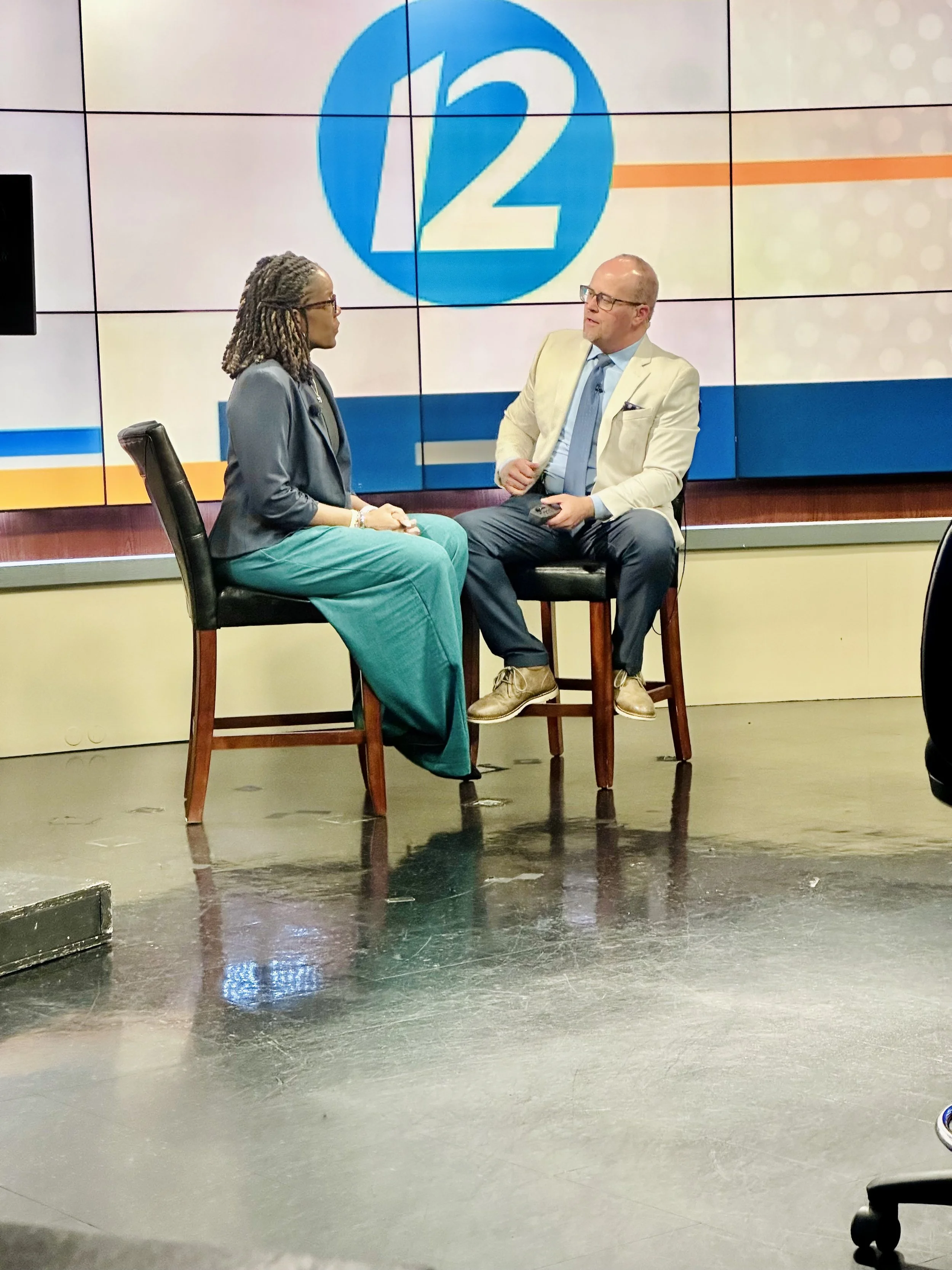 A woman and a man are sitting on chairs in a TV news studio during an interview. The woman has dark braided hair and is wearing teal pants and a dark blazer. The man is bald, wearing glasses, a light-colored blazer, light blue shirt, dark pants, tan shoes, and is holding a remote or phone. Behind them, a large screen displays a blue circle with the number 12, likely the news channel logo.