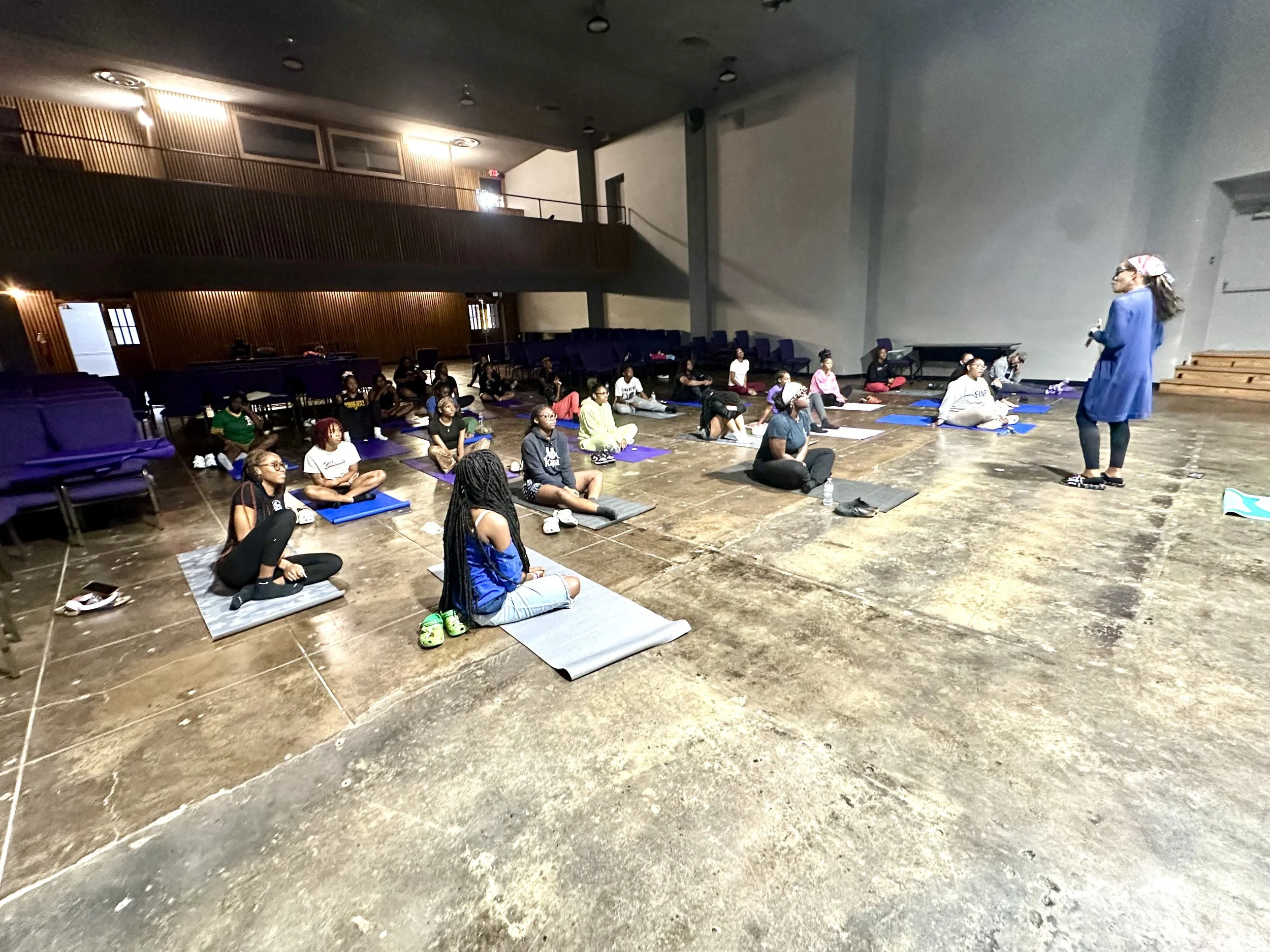 A group of people participating in a yoga or meditation class indoors, seated on mats, with an instructor standing at the front.