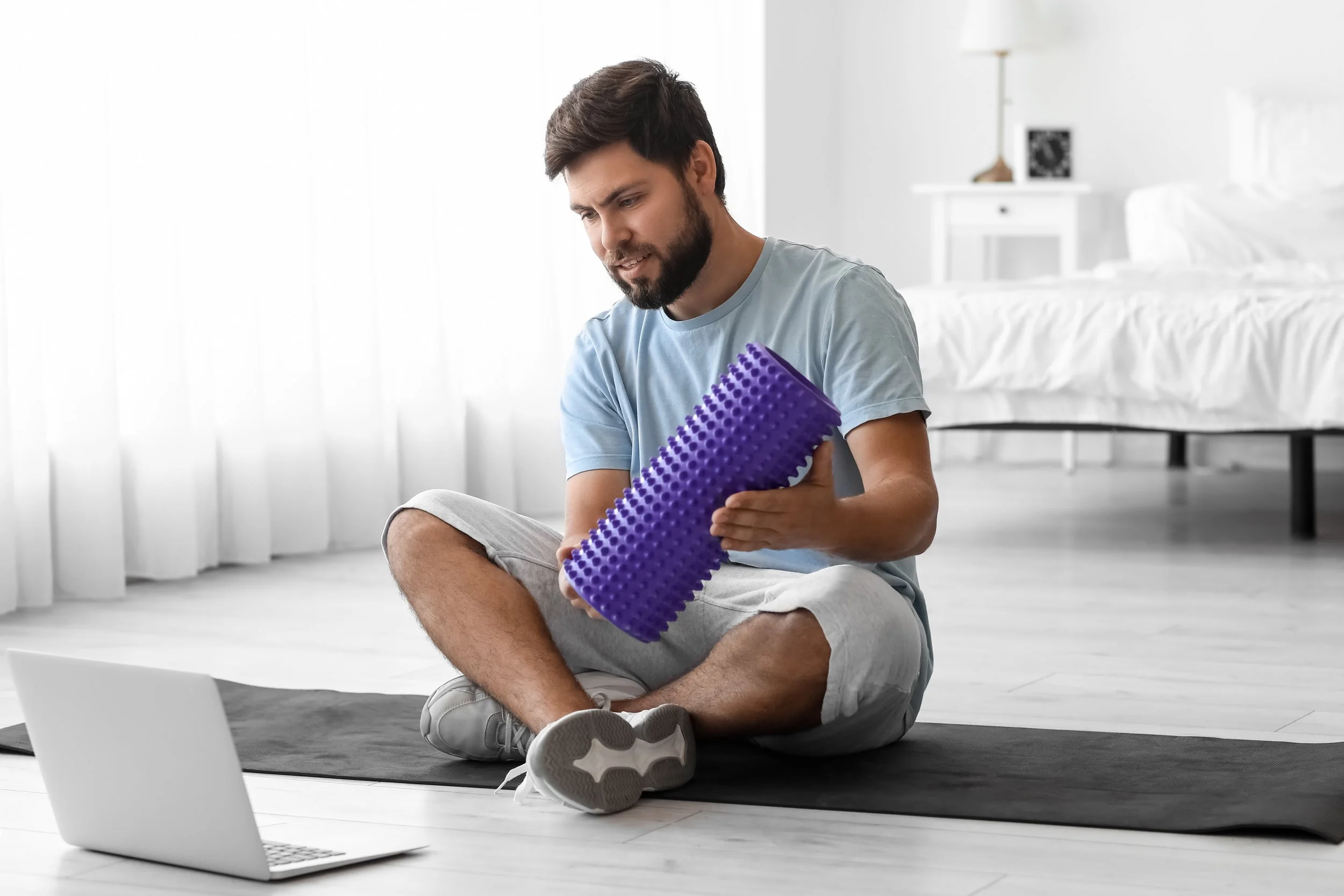 A man sitting on a yoga mat in a bright room, holding a purple foam roller and looking at a laptop placed on the floor in front of him.