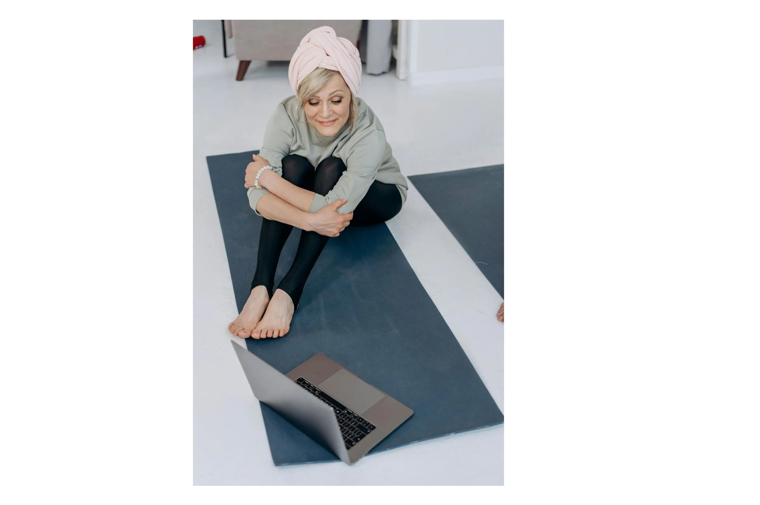 A woman with light skin, blonde hair, wearing a pink headwrap, gray top, black tights, and a pearl bracelet, sitting cross-legged on a yoga mat in a room, looking at a laptop in front of her.