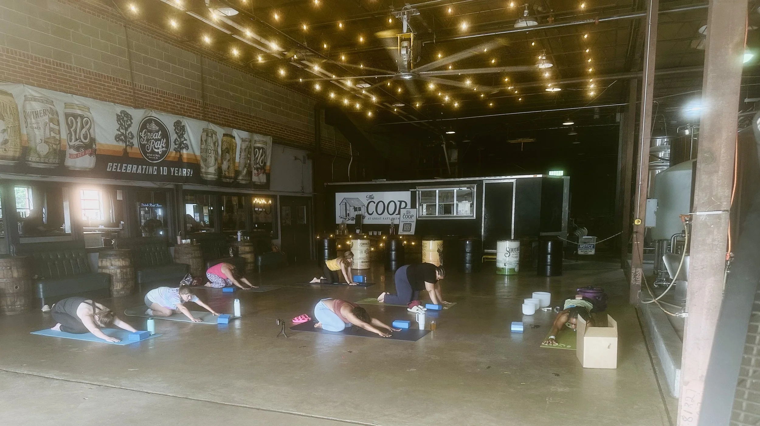 Group of people practicing yoga on mats inside a brewery, with warm lighting and industrial decor.