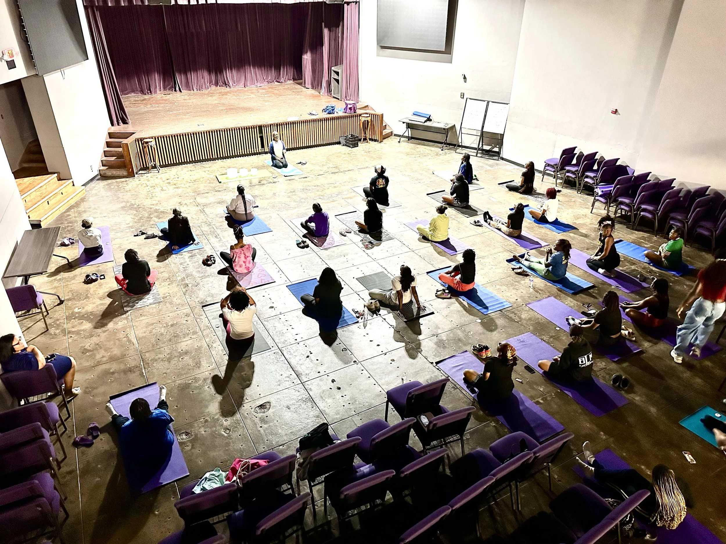 People participating in a yoga class in a large indoor room with a stage at the front, many seated on yoga mats, some seated on chairs along the sides.