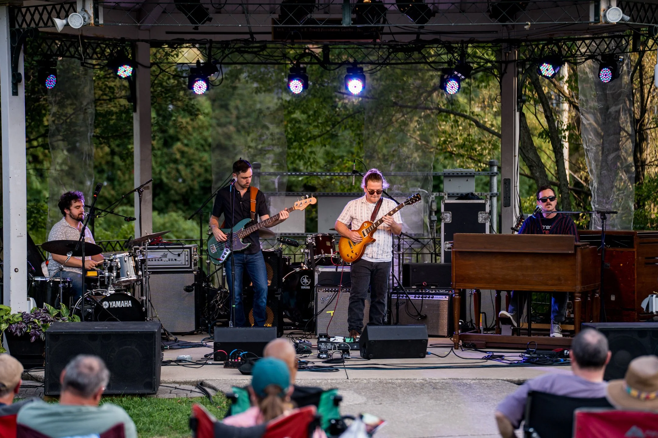 Cordelia Blue the band performing live on an outdoor stage with four musicians playing instruments, with drums, bass guitar, electric guitar, and keyboard, under stage lights during a daytime outdoor event, with an audience seated in front.