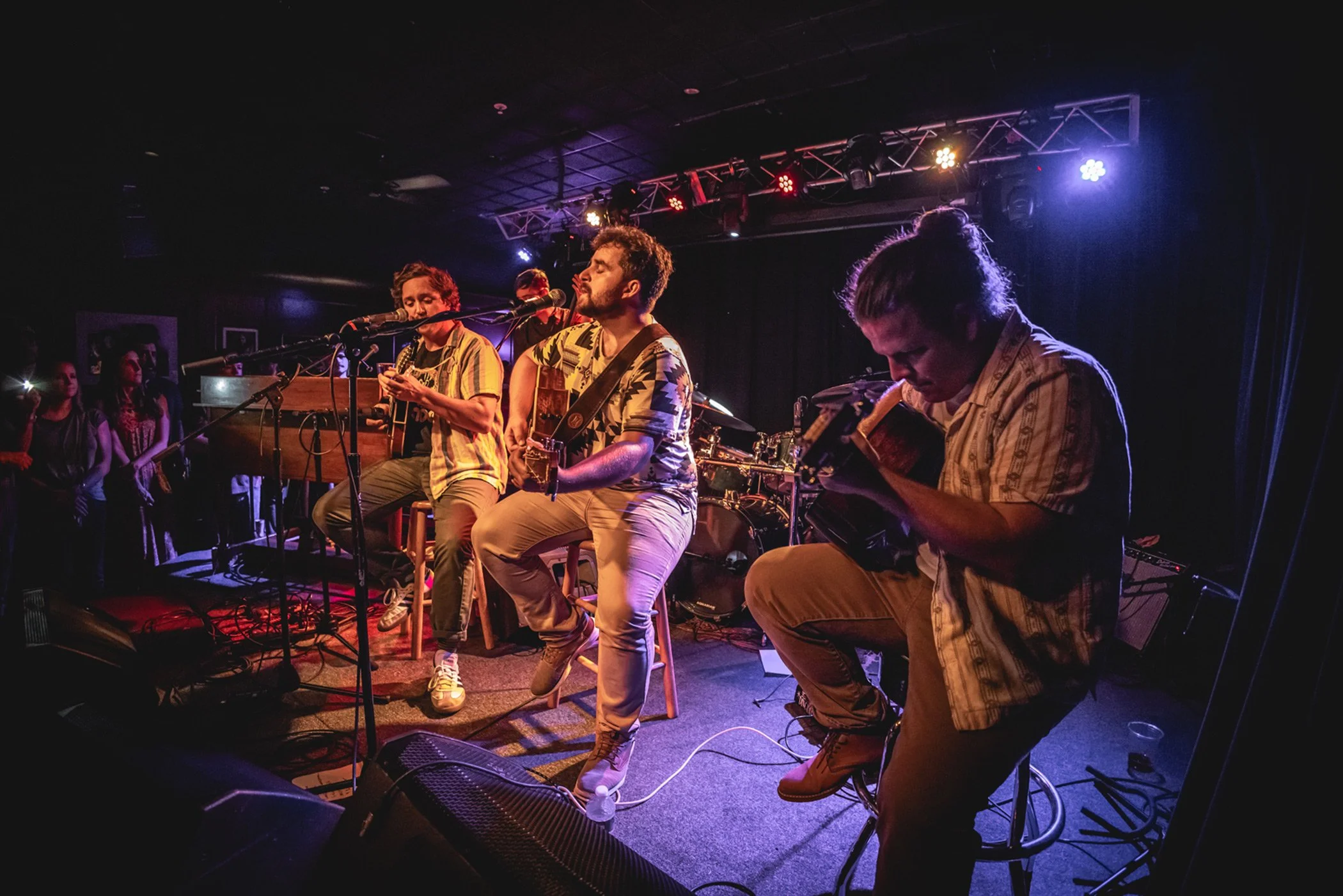 Cordelia Blue performing live on stage with four musicians playing guitars and singing into microphones, with audience members watching in the background under stage lights.