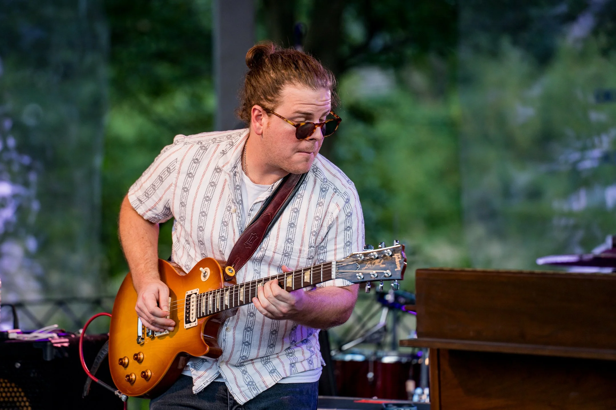 Cordelia Blue's Luke Ferracone wearing sunglasses and a striped shirt playing an electric guitar outdoors with Robert Randolph, with a blurred green background