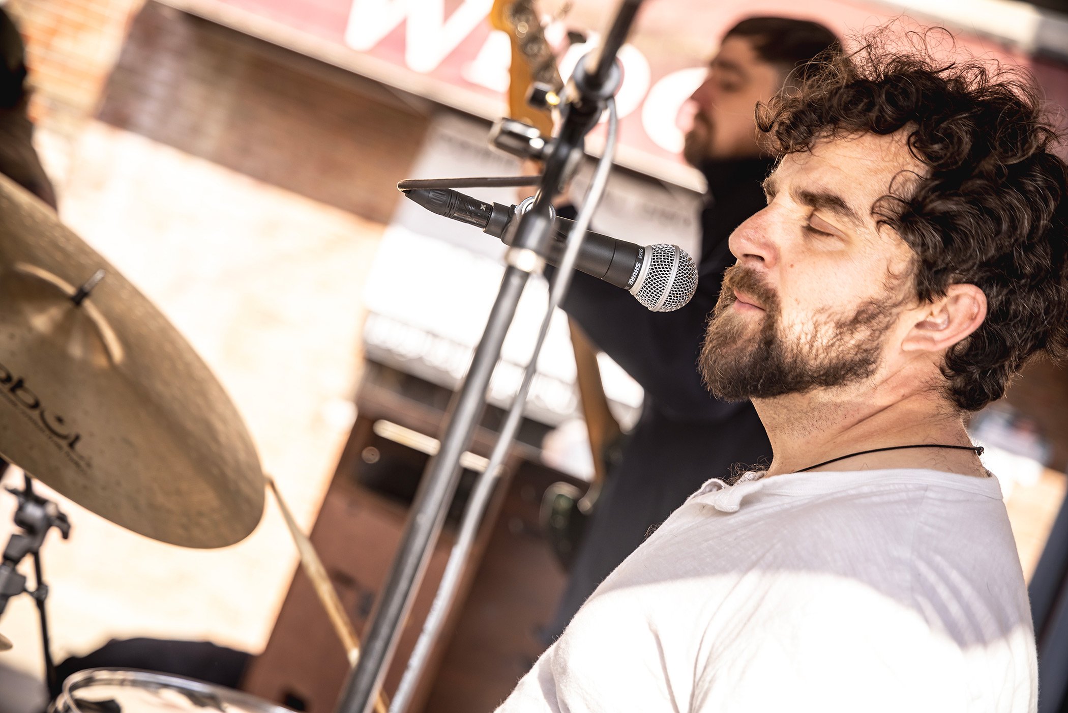 Cordelia Blue's Rob Tait sitting at a drum set in the foreground with eyes closed and the Patrick Bickel in the background smiling, both near microphones.