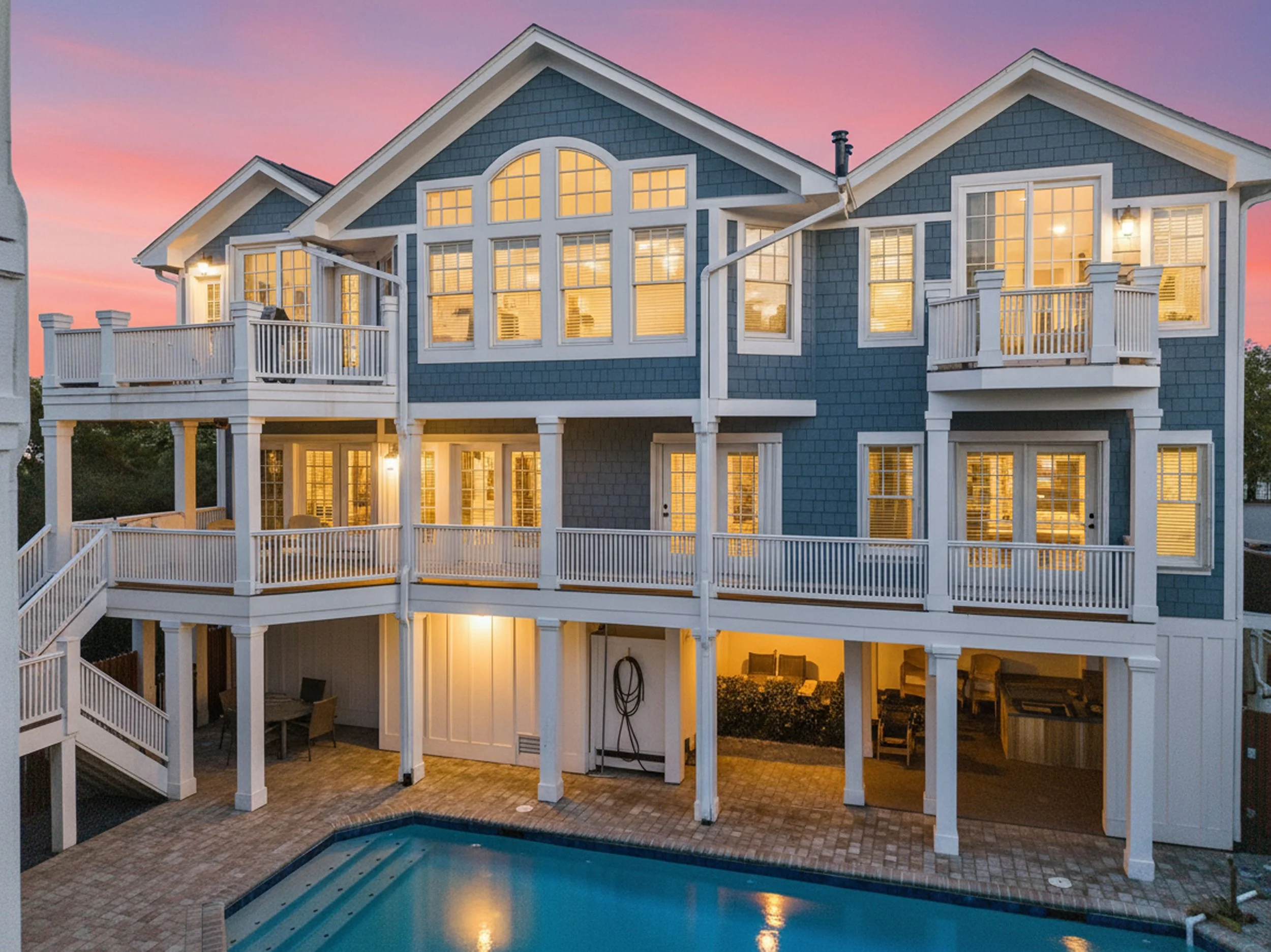 A multi-story house with blue exterior siding, white trim, multiple windows, and balconies, illuminated with interior lighting at dusk. There is also a swimming pool at the bottom of the house with patio furniture surrounding it.