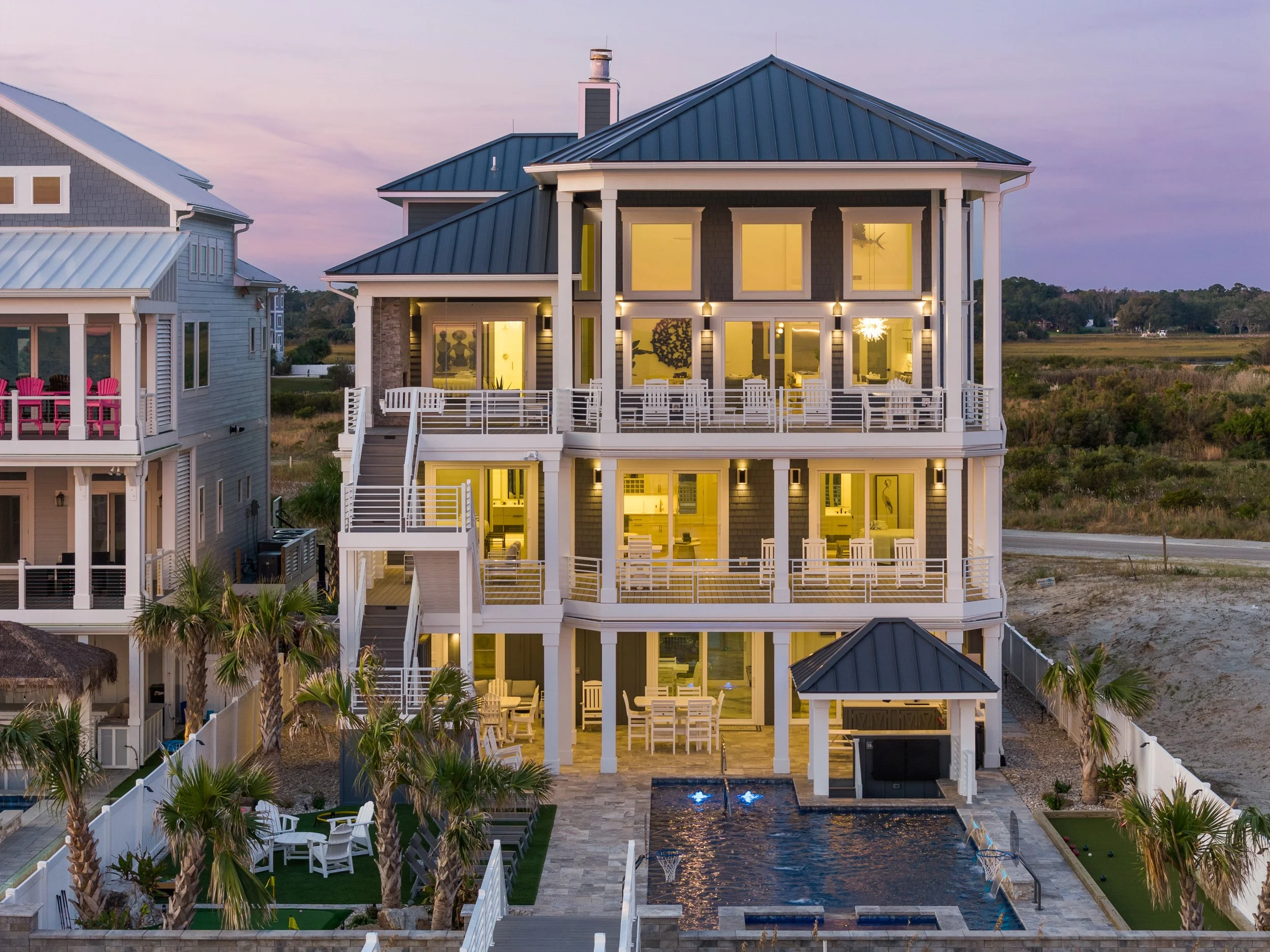 Three-story coastal house with large decks, illuminated windows, outdoor seating, swimming pool, and palm trees, during dusk.