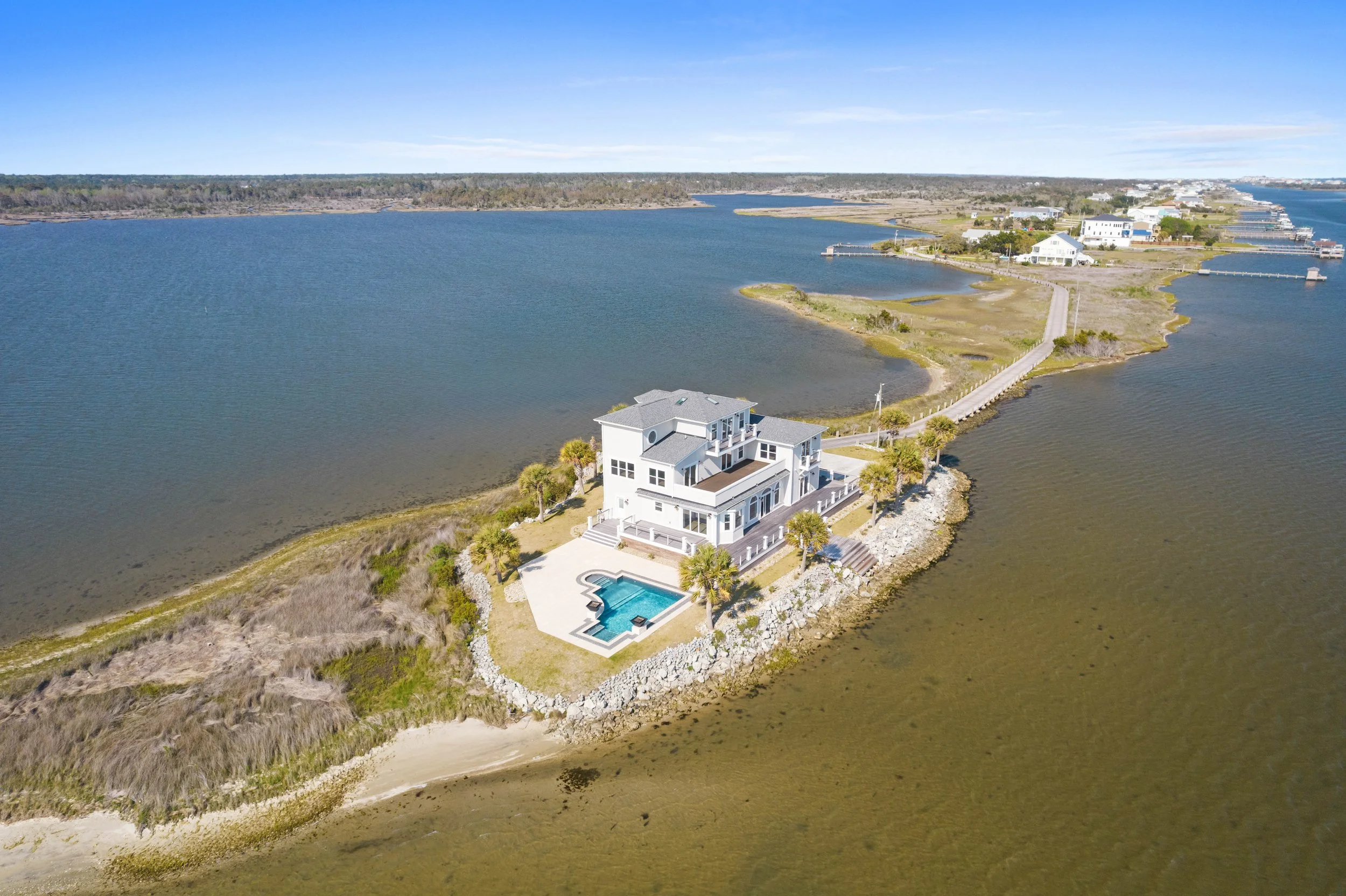 A large beach house with a swimming pool on a small island connected by a long bridge in a waterway, with a narrow sandy beach area and palm trees around the house.