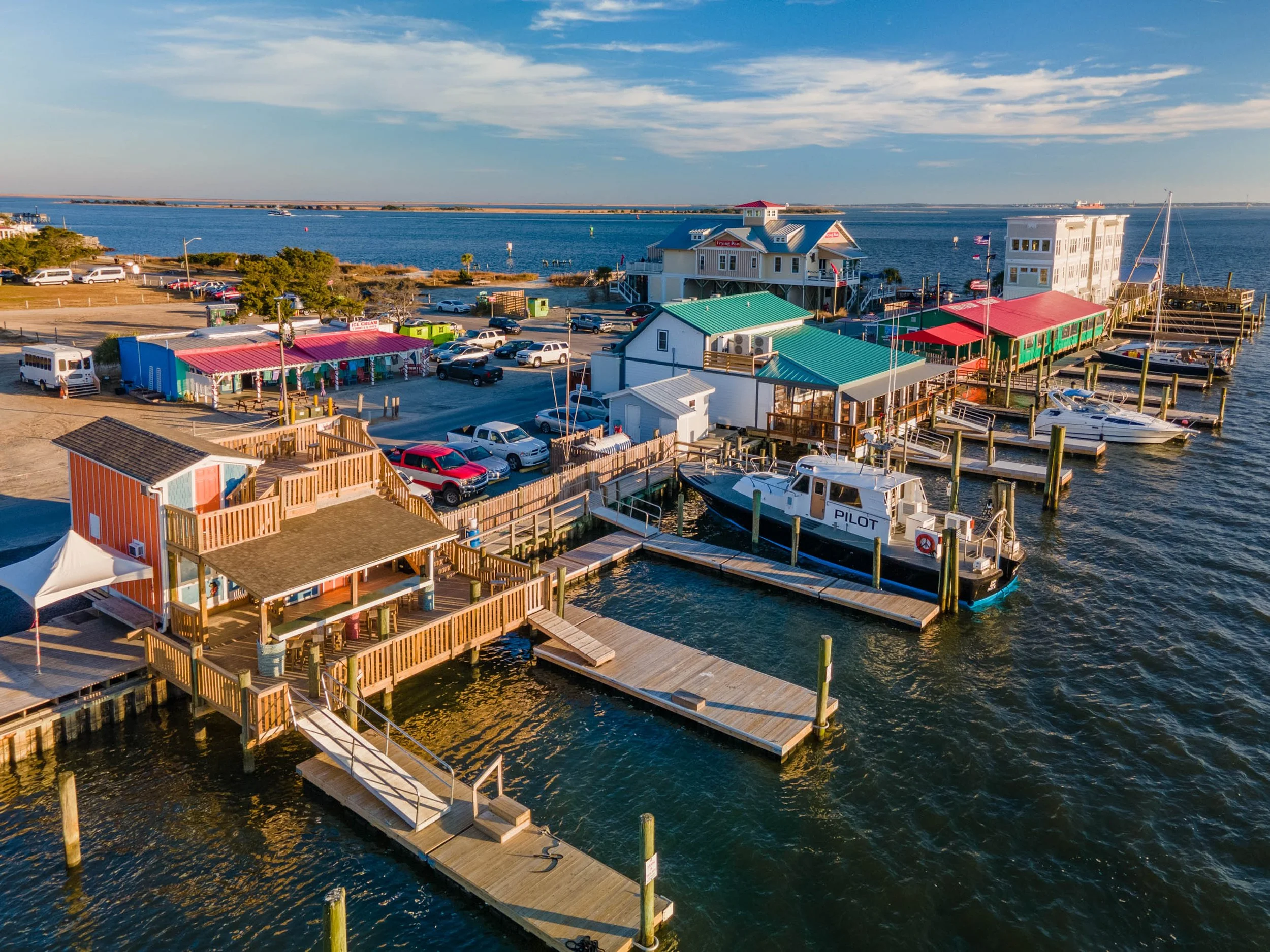 Aerial view of a small marina with boats docked, colorful buildings, parked cars, and a waterfront with water and a distant harbor under a blue sky.