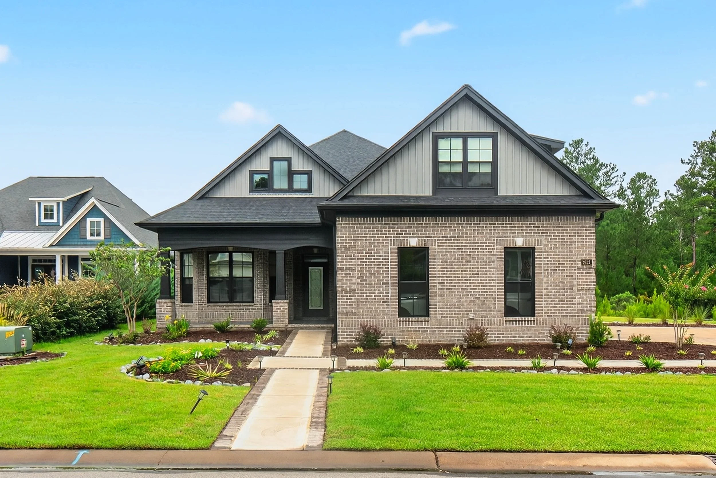 Front view of a modern two-story house with a brick and gray siding exterior, surrounded by a green lawn and landscaped yard, under a blue sky with a few clouds.