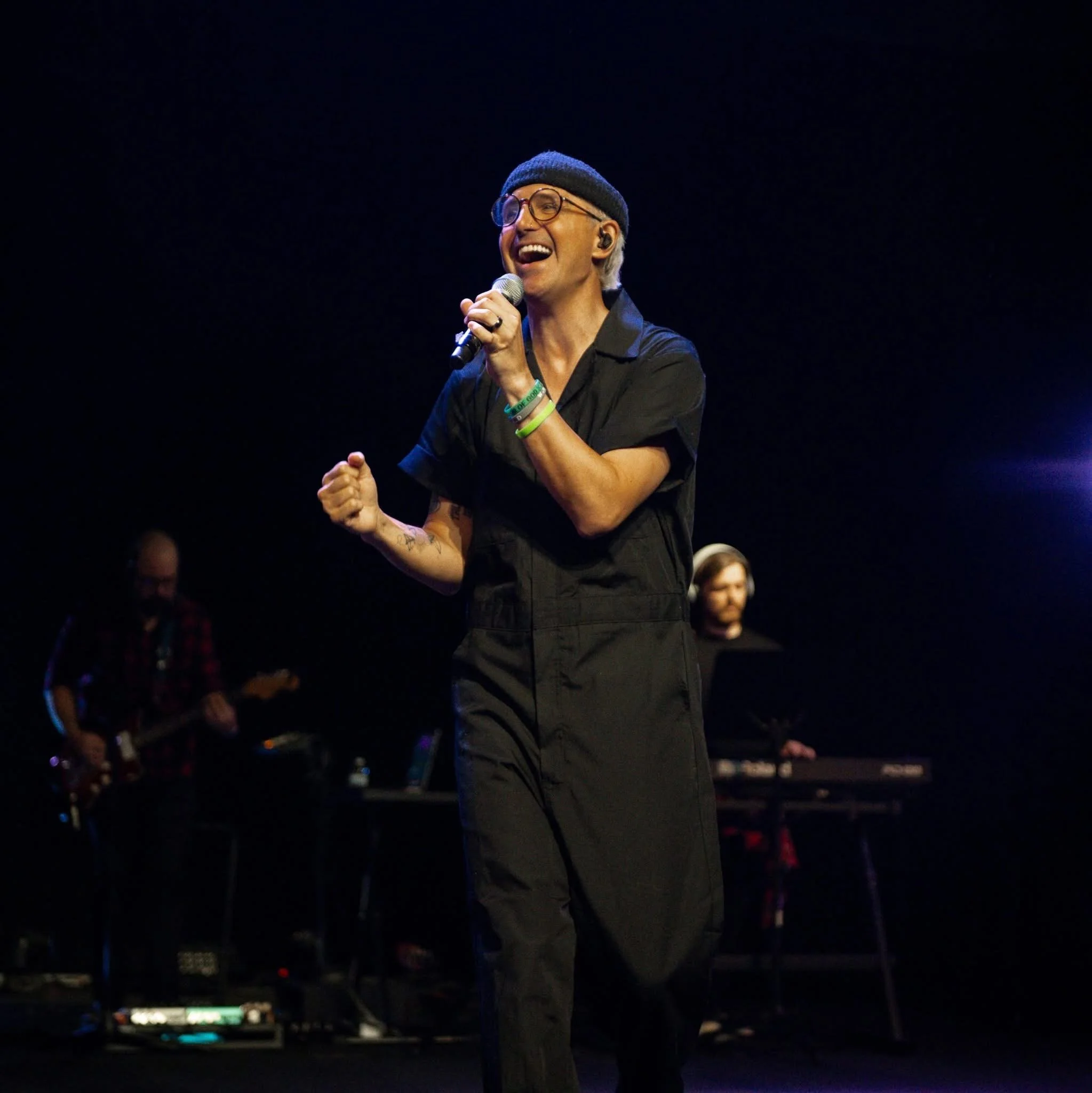 A person performing on stage, wearing glasses, a beanie, and a black shirt, holding a microphone, with a dark background and some audience members visible.