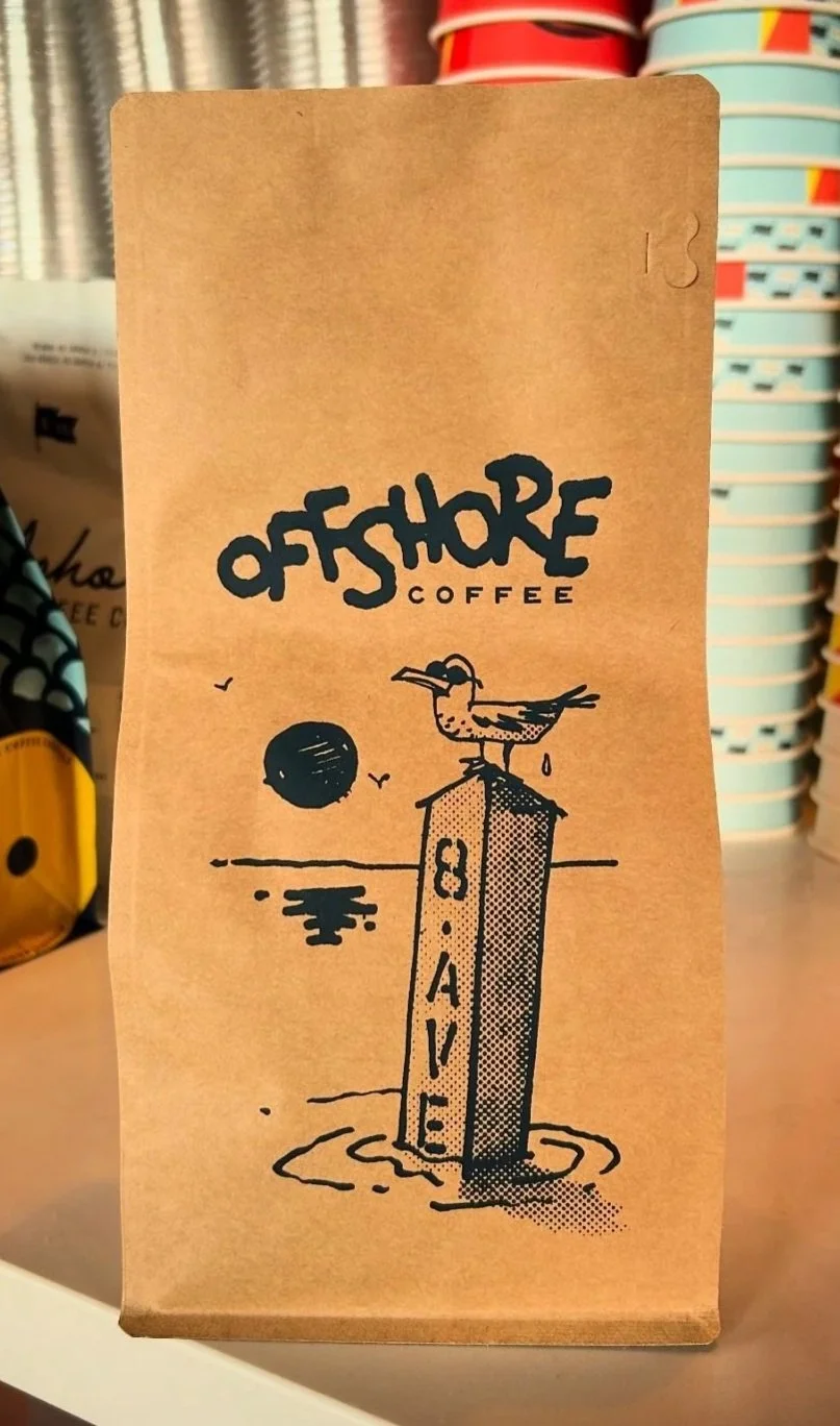 A brown paper coffee bag from Off Shore Coffee decorated with a black illustration of a bird standing on a pole marked 'Beach' near water with a setting sun in the background.