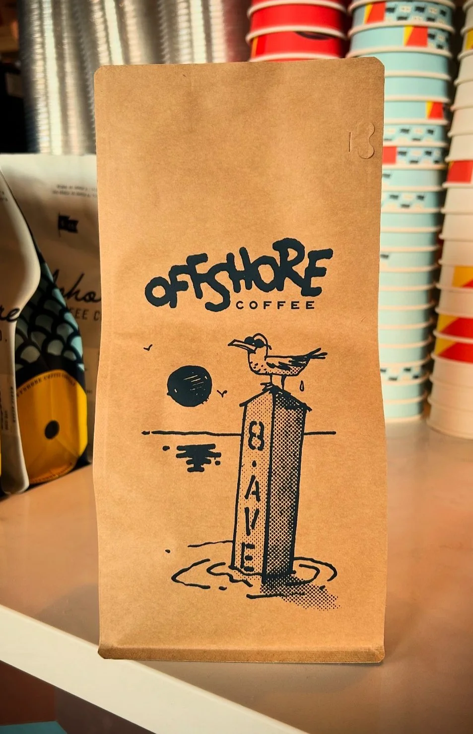 A coffee bag from Offshore Coffee featuring a graphic of a seagull standing on a signpost marked 'Beach,' with the sun setting over water in the background and a few seagulls flying.