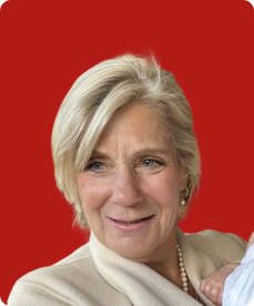 A close-up of a smiling woman with short, blonde hair, wearing a cream-colored top and pearl necklace, against a solid red background.