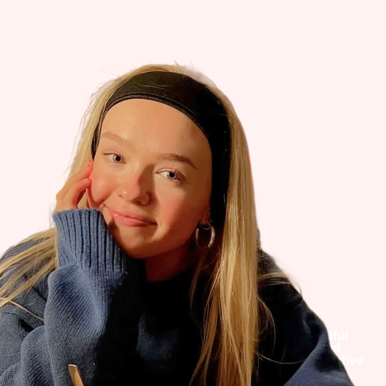 A young woman with long blonde hair, wearing a black headband and a blue and black hoodie, resting her face on her hand and smiling softly.
