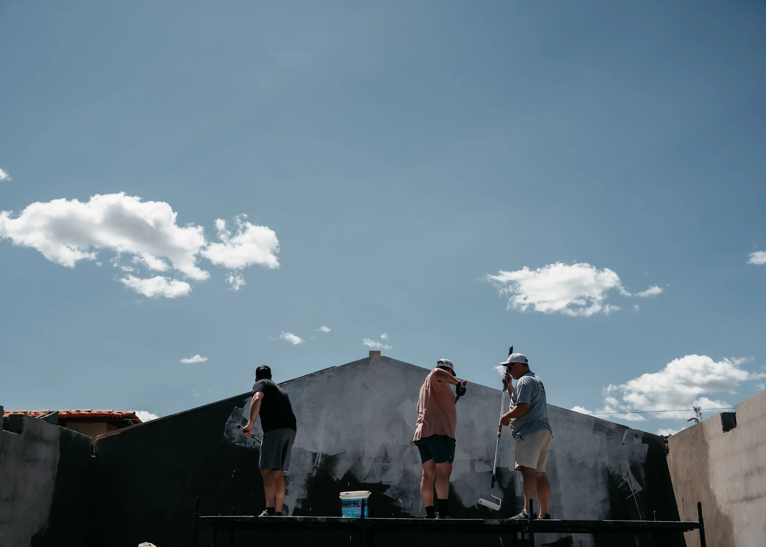 Three people painting a wall outdoors with a blue sky and clouds above.