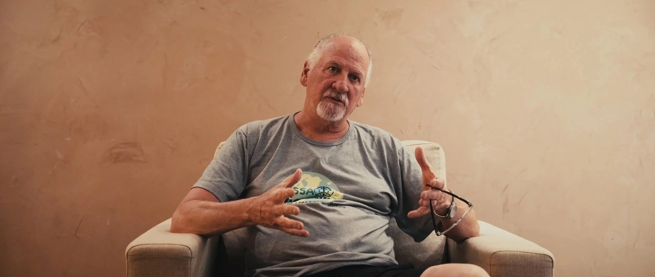 An older man with a beard and glasses, wearing a gray T-shirt, sitting in a beige armchair against a beige wall, making hand gestures.