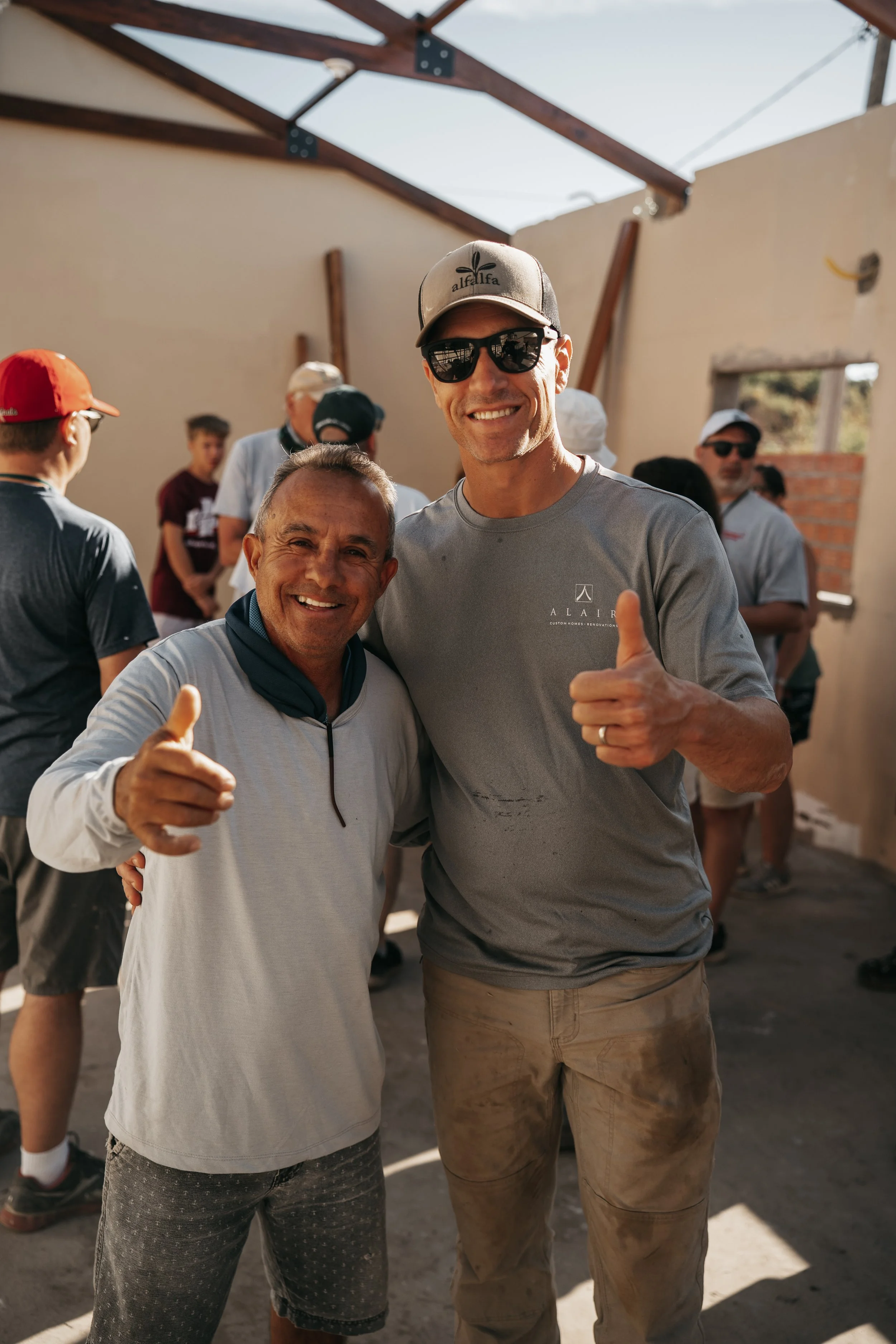 Two men smiling and giving thumbs up at an indoor construction site event, with several people in the background.