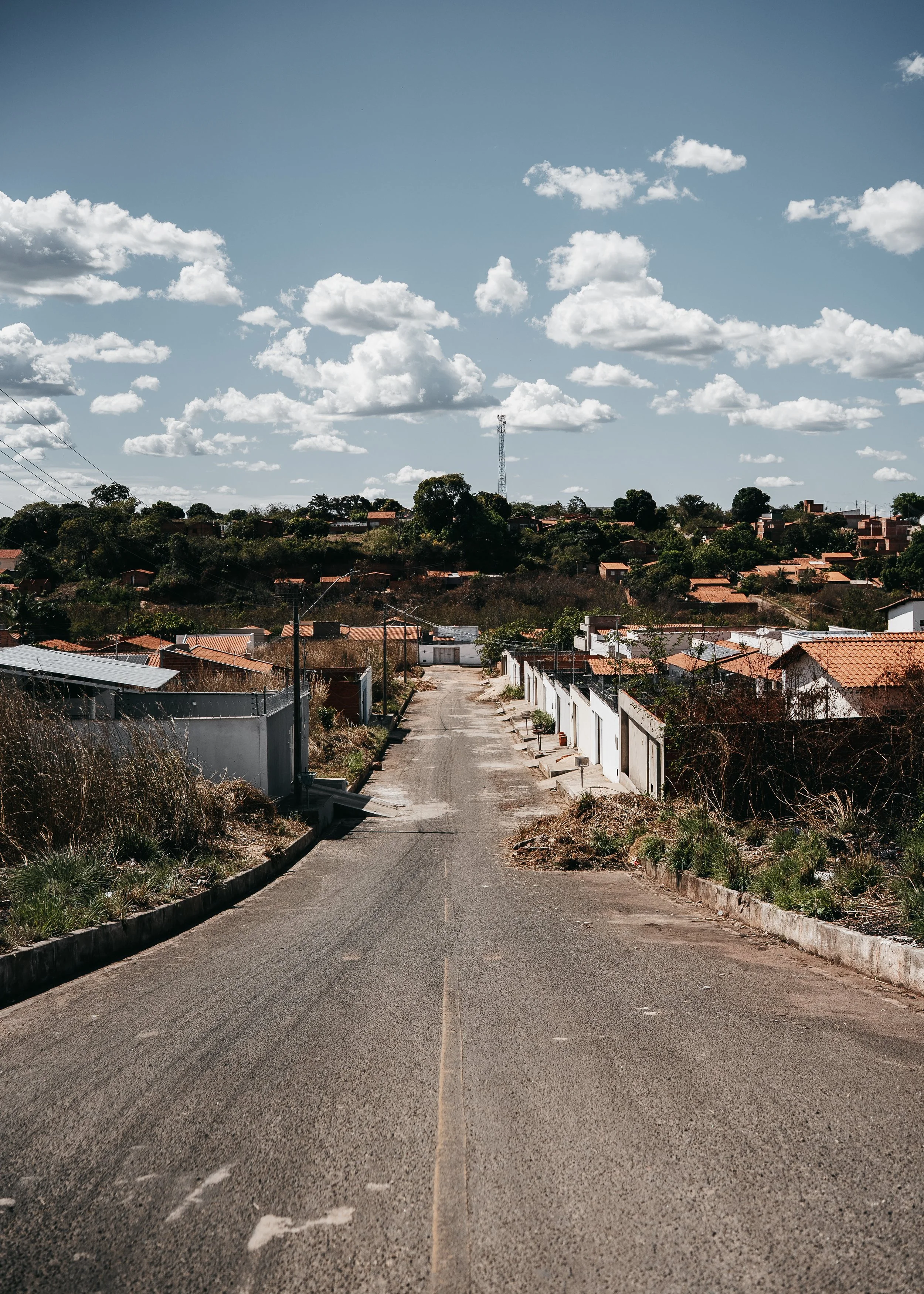 A quiet, sloped residential street with a single lane, bordered by fences and dry grass, leading to a hilly neighborhood with houses and trees under a partly cloudy sky.