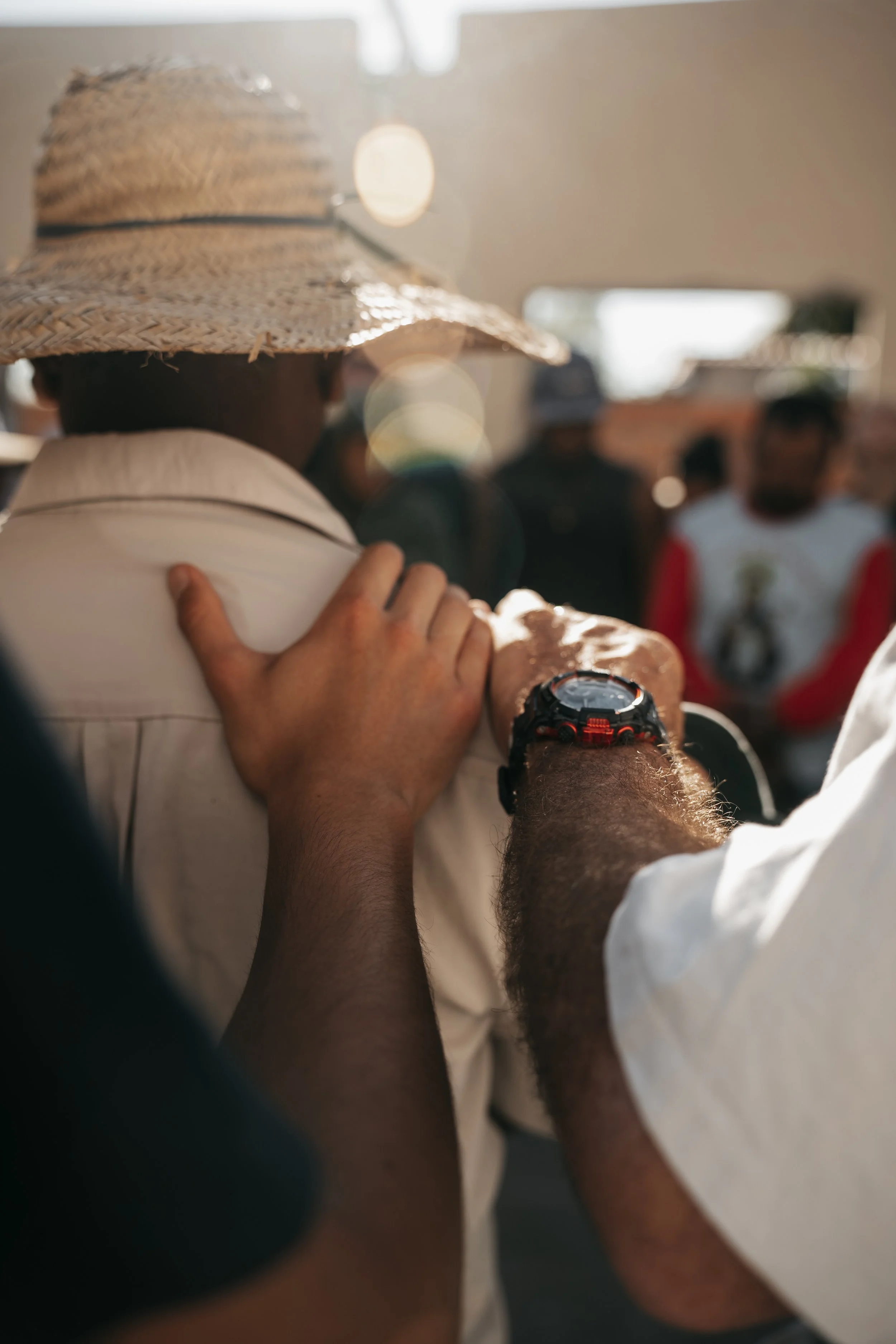 A person wearing a straw hat and a white shirt is holding another person's shoulder. The scene is outdoors with blurred people in the background.
