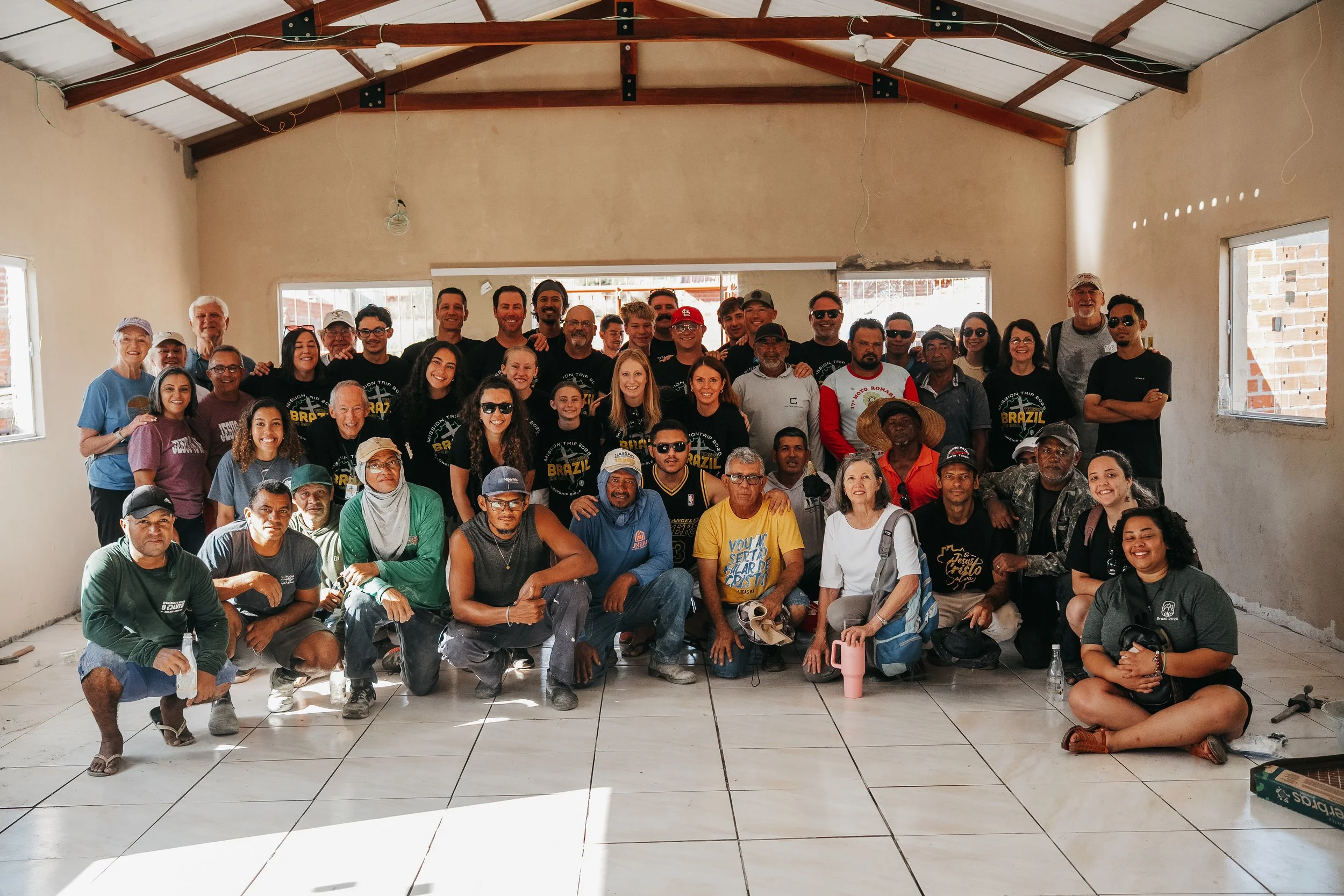 Group of diverse people gathered in a partially constructed or renovated room with unfinished walls and a high ceiling, posing for a photo, some smiling and wearing casual clothing.