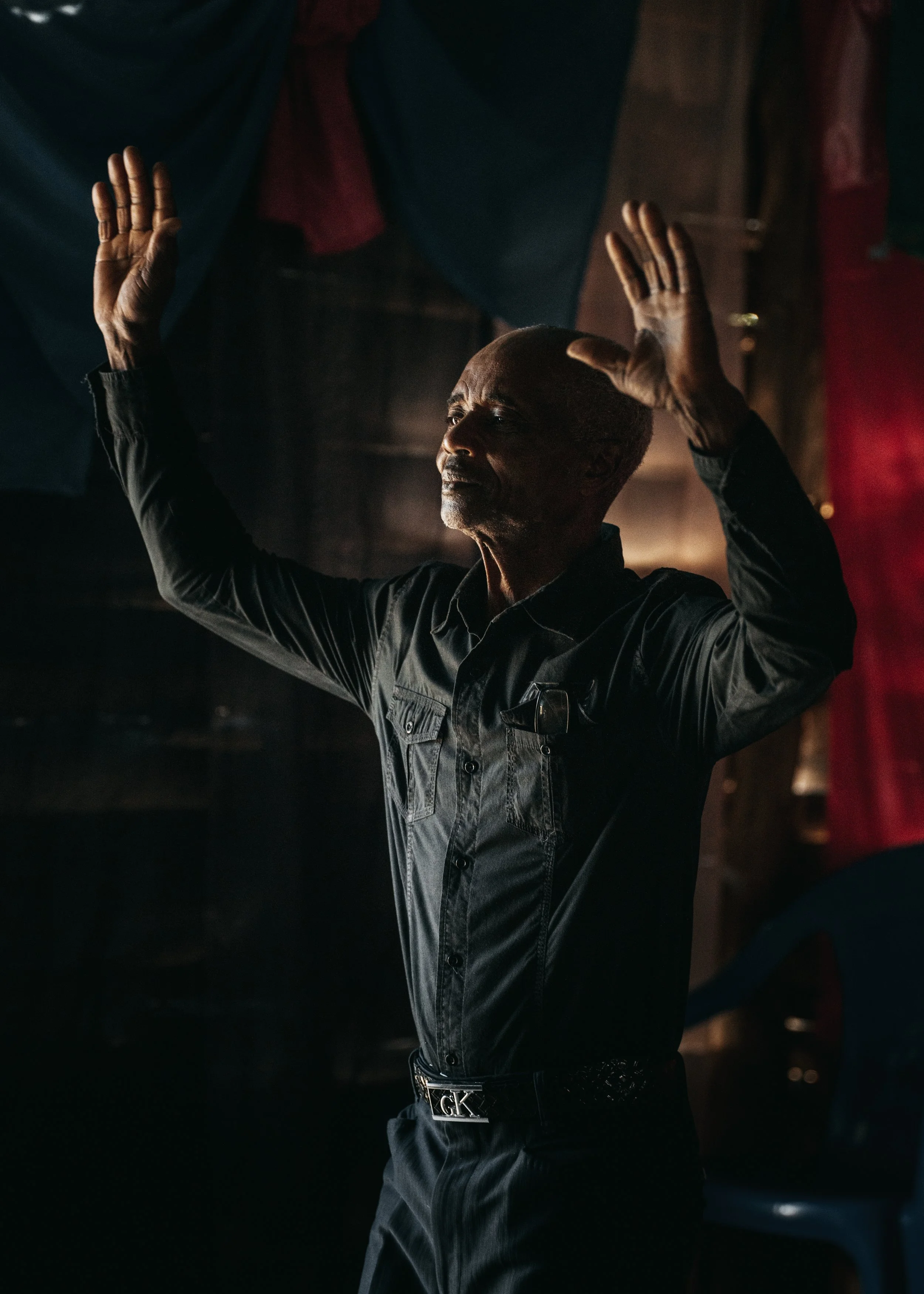 A man with a shaved head and gray beard raising both hands with palms facing outward in a dark indoor setting.