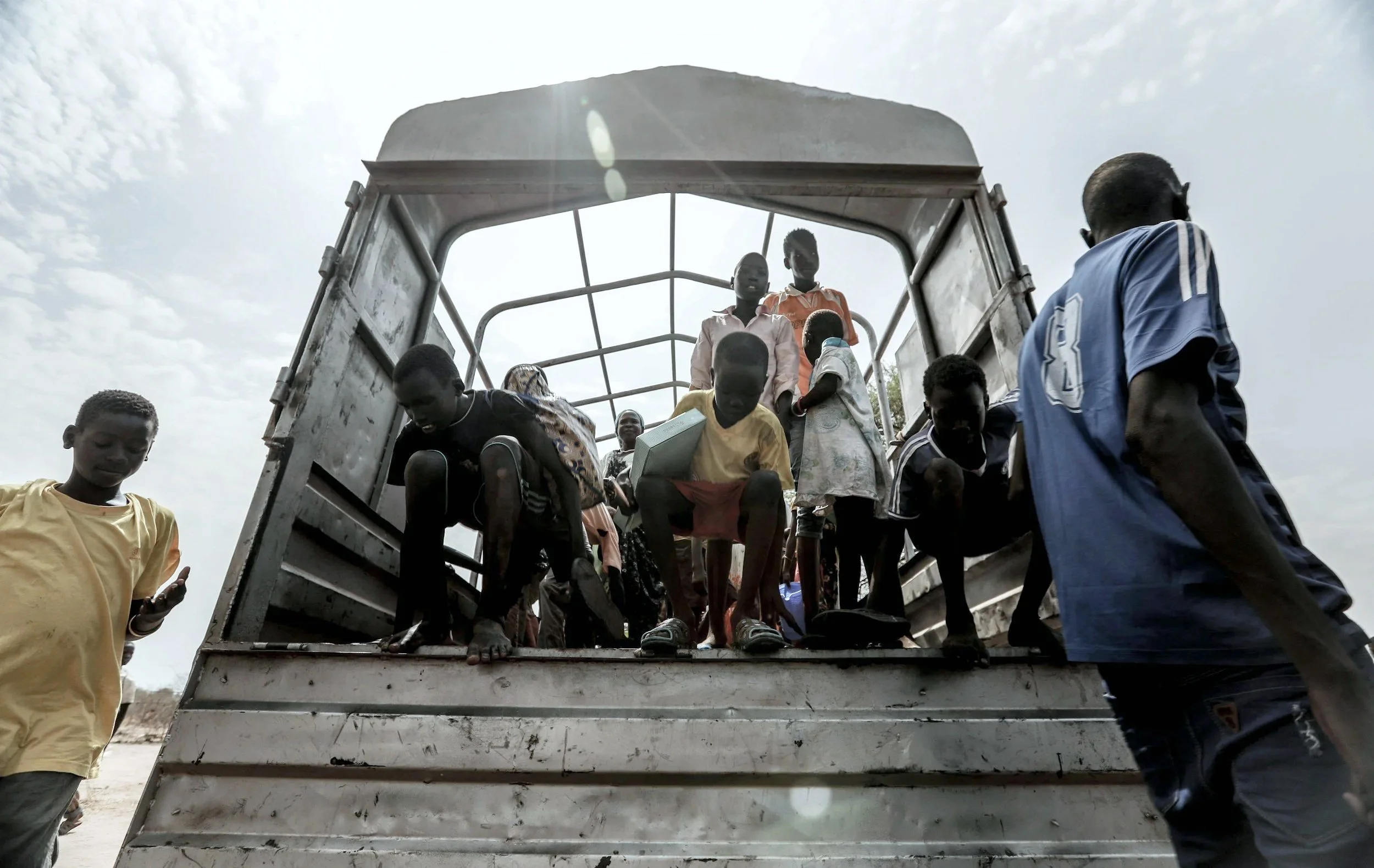 Children and adults boarding a truck, possibly in a refugee or displaced persons scenario, under a cloudy sky.