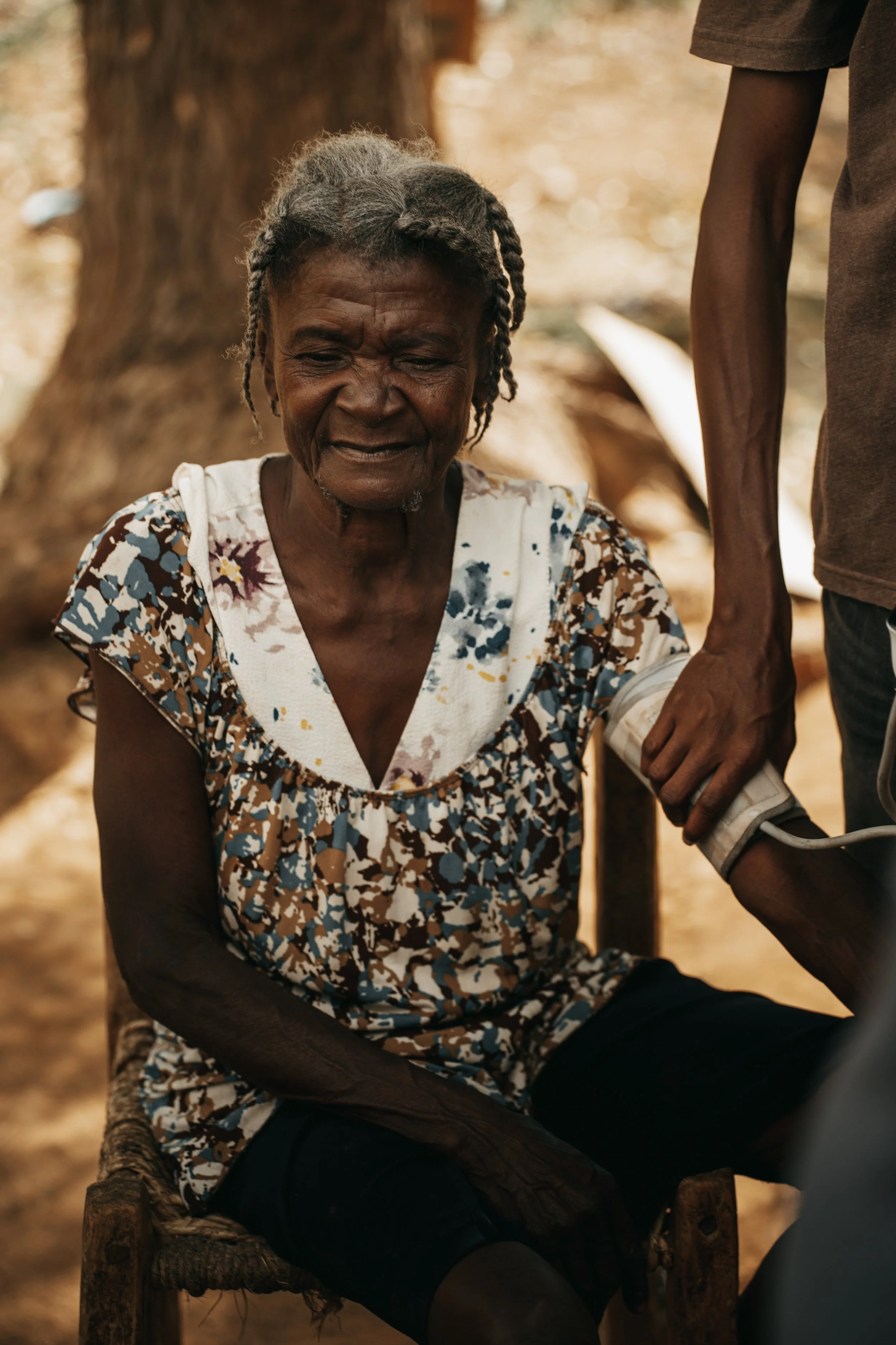 An elderly woman sitting outdoors having her blood pressure checked by a person standing next to her.