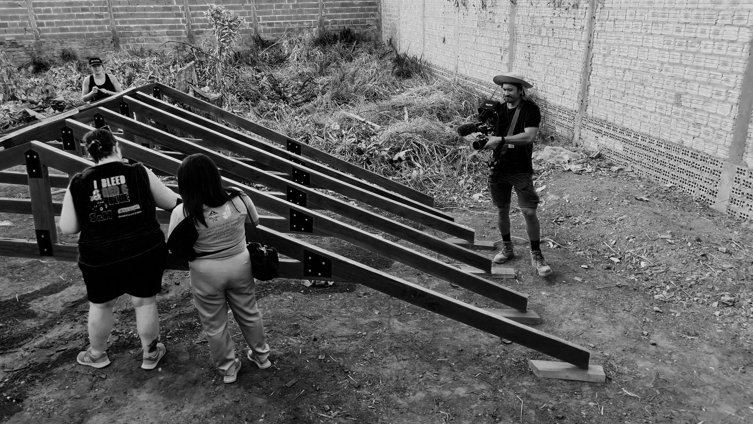 A film crew filming two women and a girl at an outdoor construction site with stairs and debris, with a man holding a camera and wearing a sombrero.