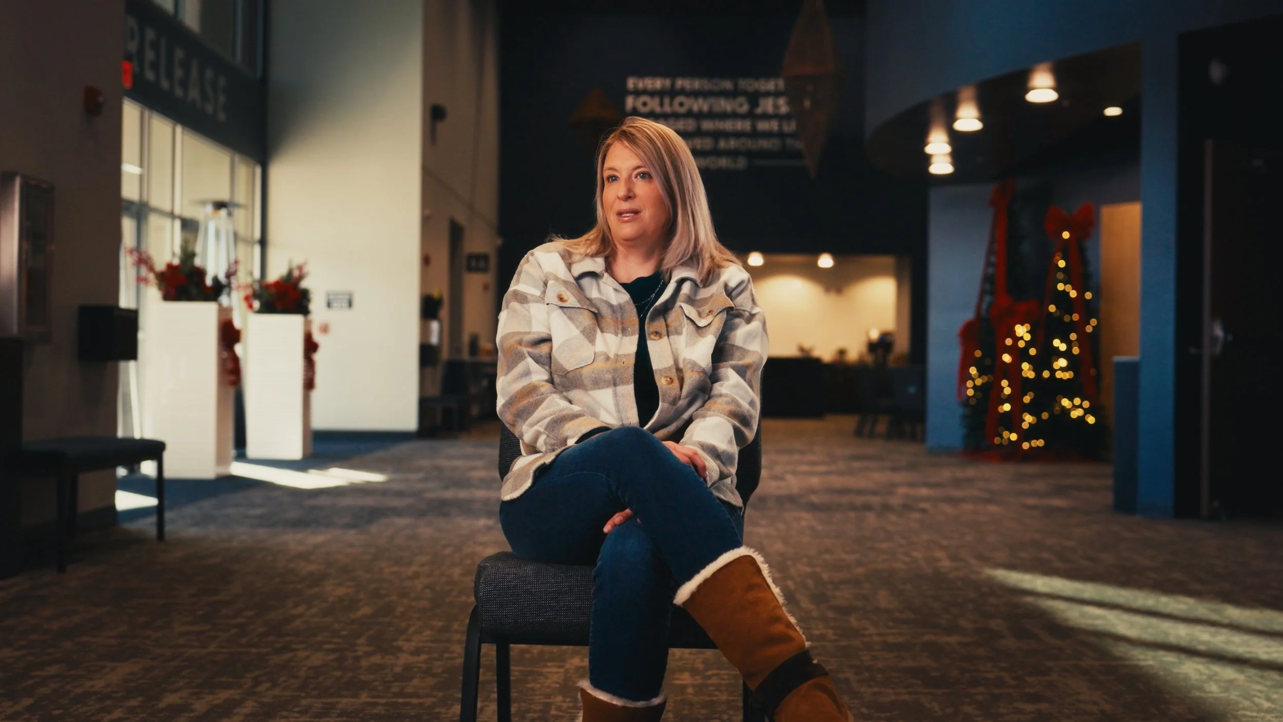 A woman sitting on a chair in a lobby decorated with Christmas decorations, including a Christmas tree with lights in the background.