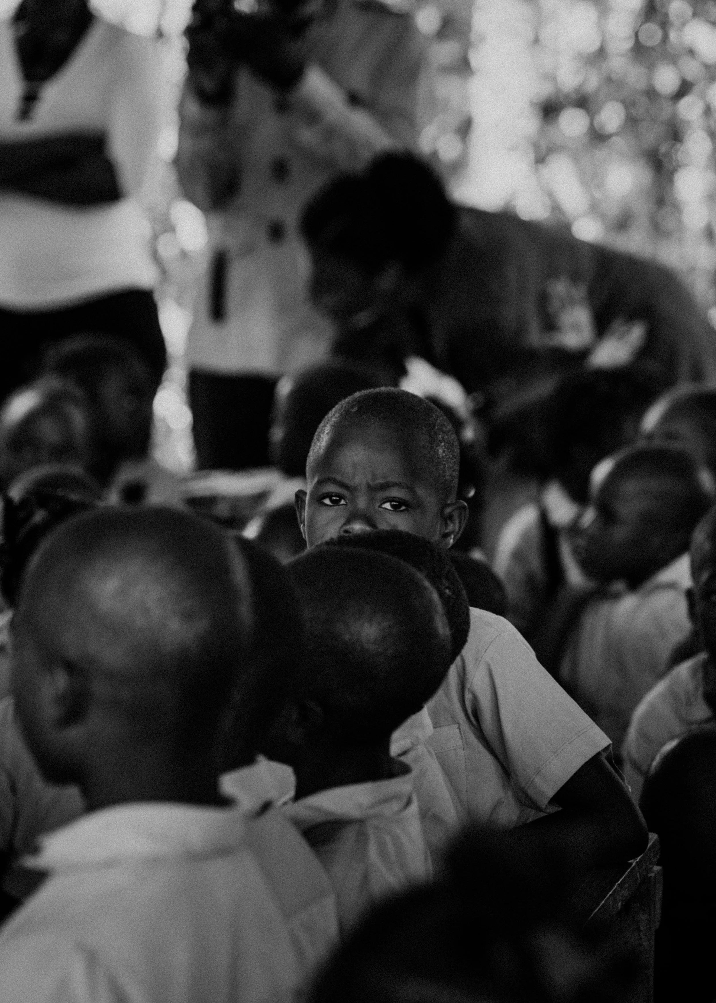 A black and white photo of a group of children, with one boy in the center looking directly at the camera, surrounded by other children with their heads turned or looking away.
