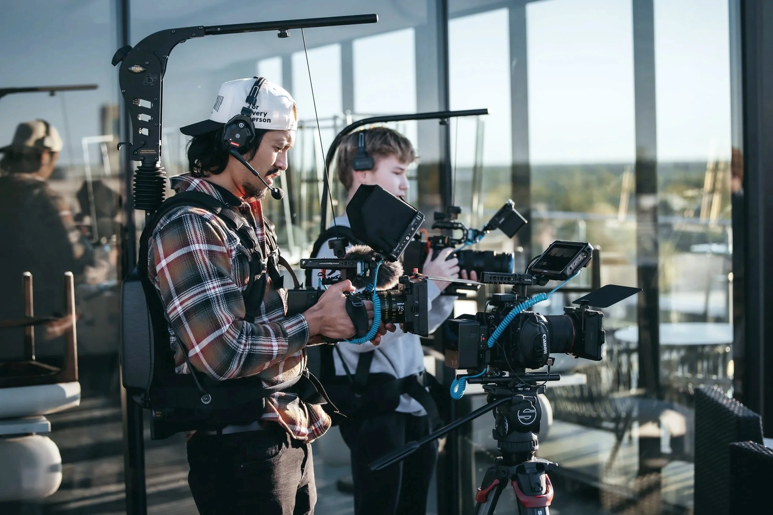 Two filmmakers working on a production set inside a building with large windows, using professional cameras and equipment.