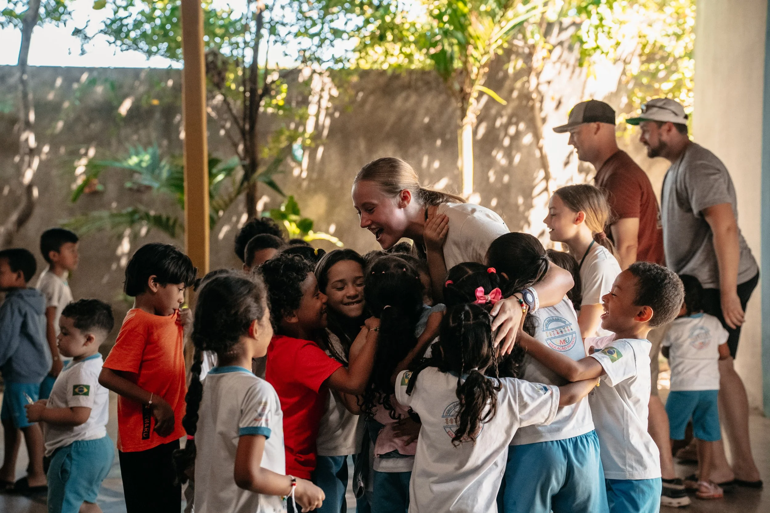 A group of children hugging a young woman in a cheerful, outdoor setting with sunlight filtering through trees.